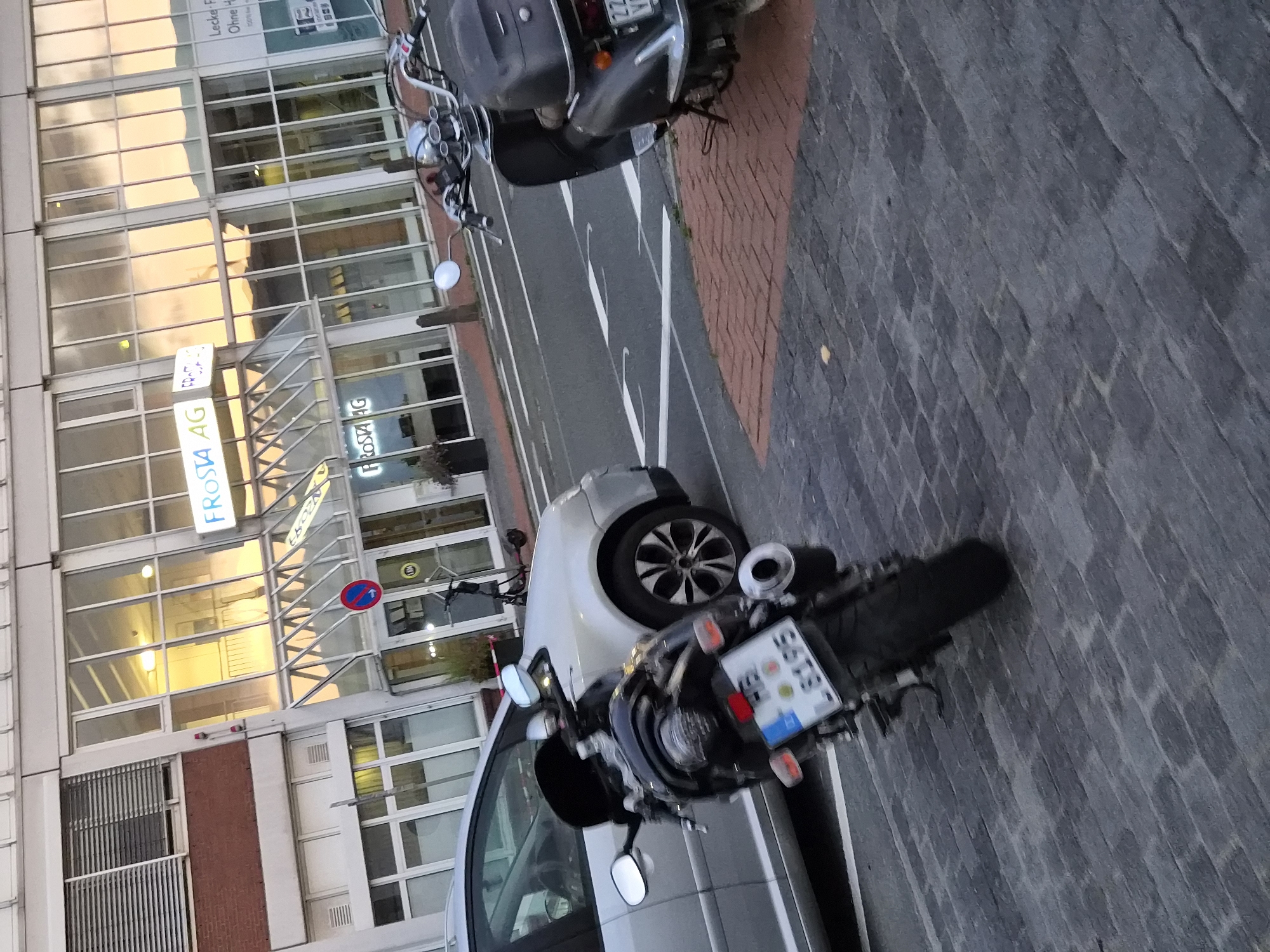 The image shows a street scene in Bremerhaven, Germany, featuring a building with large glass windows and the signage "FROSTA AG" visible. In the foreground, a black motorcycle with a license plate reading "LB195" is parked on a paved surface, with a silver car to its left. A dark-colored scooter is parked to the right, behind the motorcycle. Traffic markings are visible on the street. The lighting suggests it is daytime, with reflections of the sky in the building's windows.