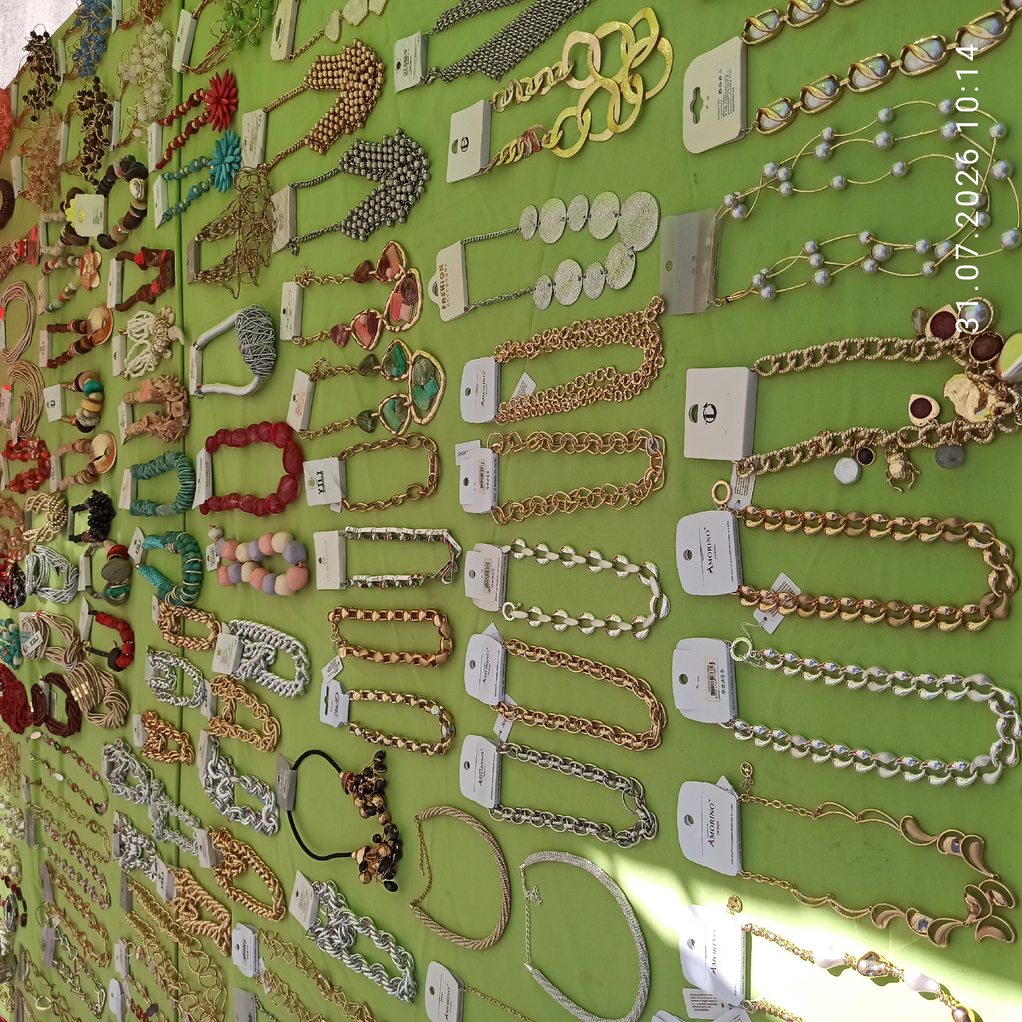A street market stall in Modugno, Italy, displays a variety of necklaces on a green tablecloth. The necklaces are arranged in rows and include various styles: chunky chains, beaded strands, pendants with colorful stones, and intricately woven designs. Some necklaces are displayed with their price tags and branding. The overall impression is one of a vibrant marketplace offering fashion accessories. The timestamp 31.07.2026 10:14 is superimposed on the image.