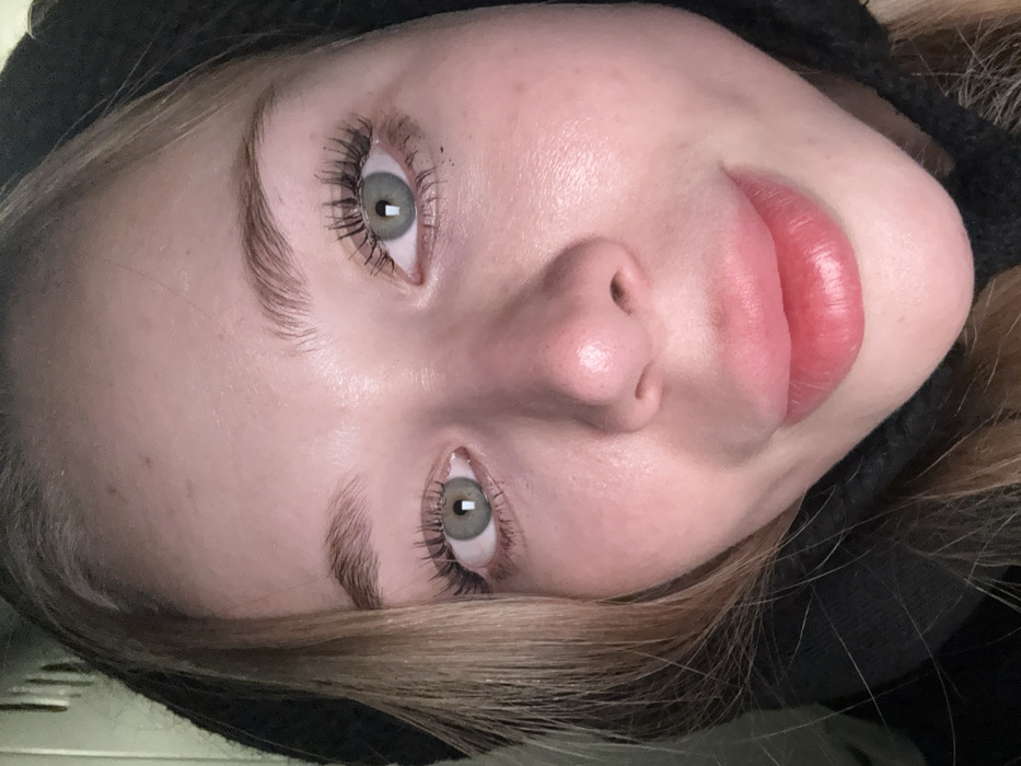 A close-up captures a fair-skinned person with striking gray-green eyes and full, glossy lips. Dark mascara emphasizes long eyelashes. Softly defined eyebrows frame the face. The individual is wearing a dark hoodie, and blonde hair is visible at the edges. The lighting is diffused, creating a soft focus on the skin. The image is oriented at an angle, presenting a unique perspective of the subject.