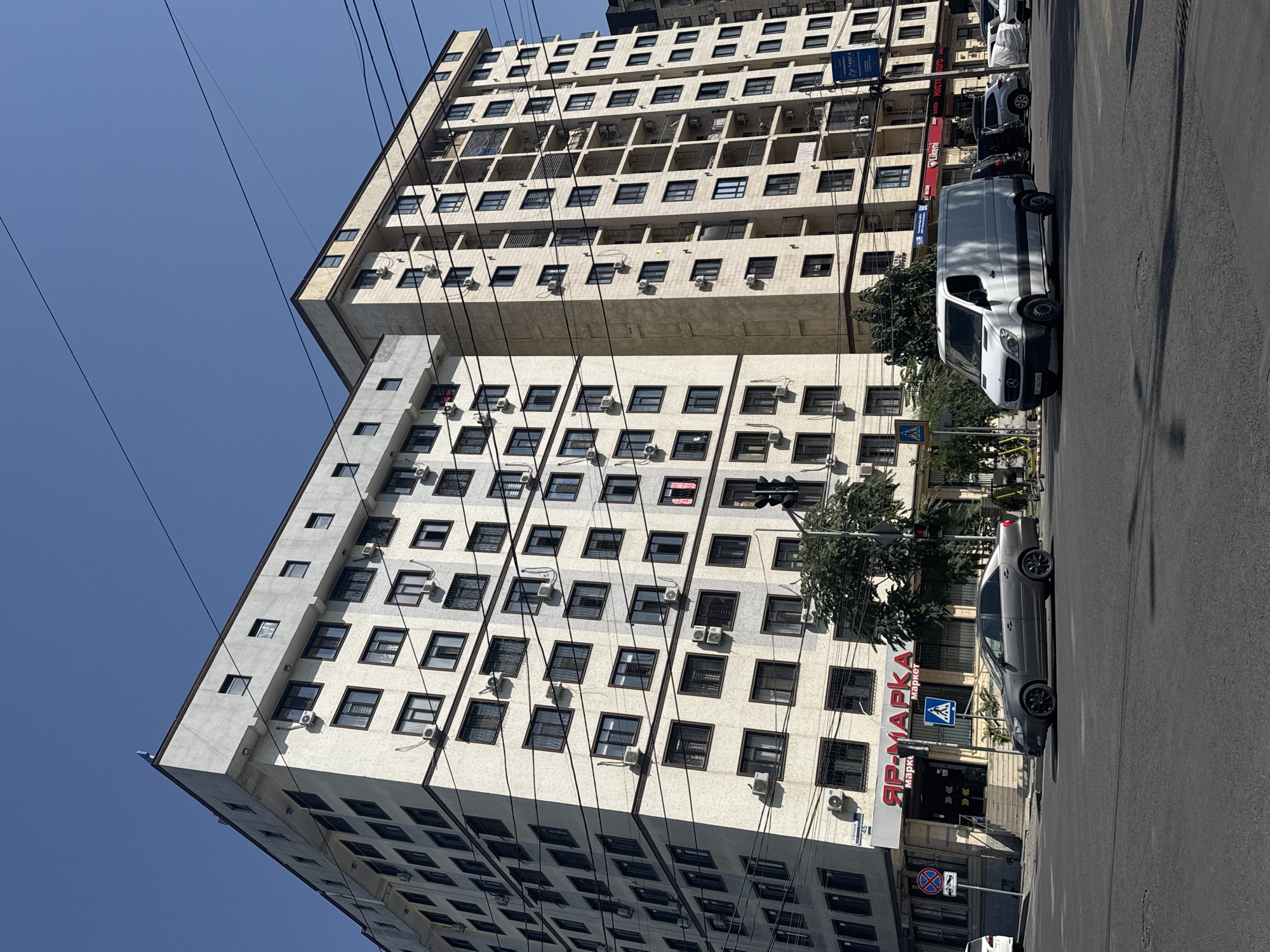 This is a street-level, eye-level, medium shot taken on a sunny day in Bishkek, Kyrgyzstan. The image features two multi-story apartment buildings with light-colored facades, many windows, and numerous external air conditioning units. The buildings are situated at a street intersection. In the foreground, a grey sedan is parked on the street, and a white van is driving past. A street sign with the number "45" is visible on the corner building. A supermarket with a red and white sign reading "ЯР-МАРКА" is located on the ground floor of one of the buildings. Traffic lights and pedestrian crossing signs are also present. Overhead power lines are visible against the clear blue sky. The scene depicts a typical urban environment.