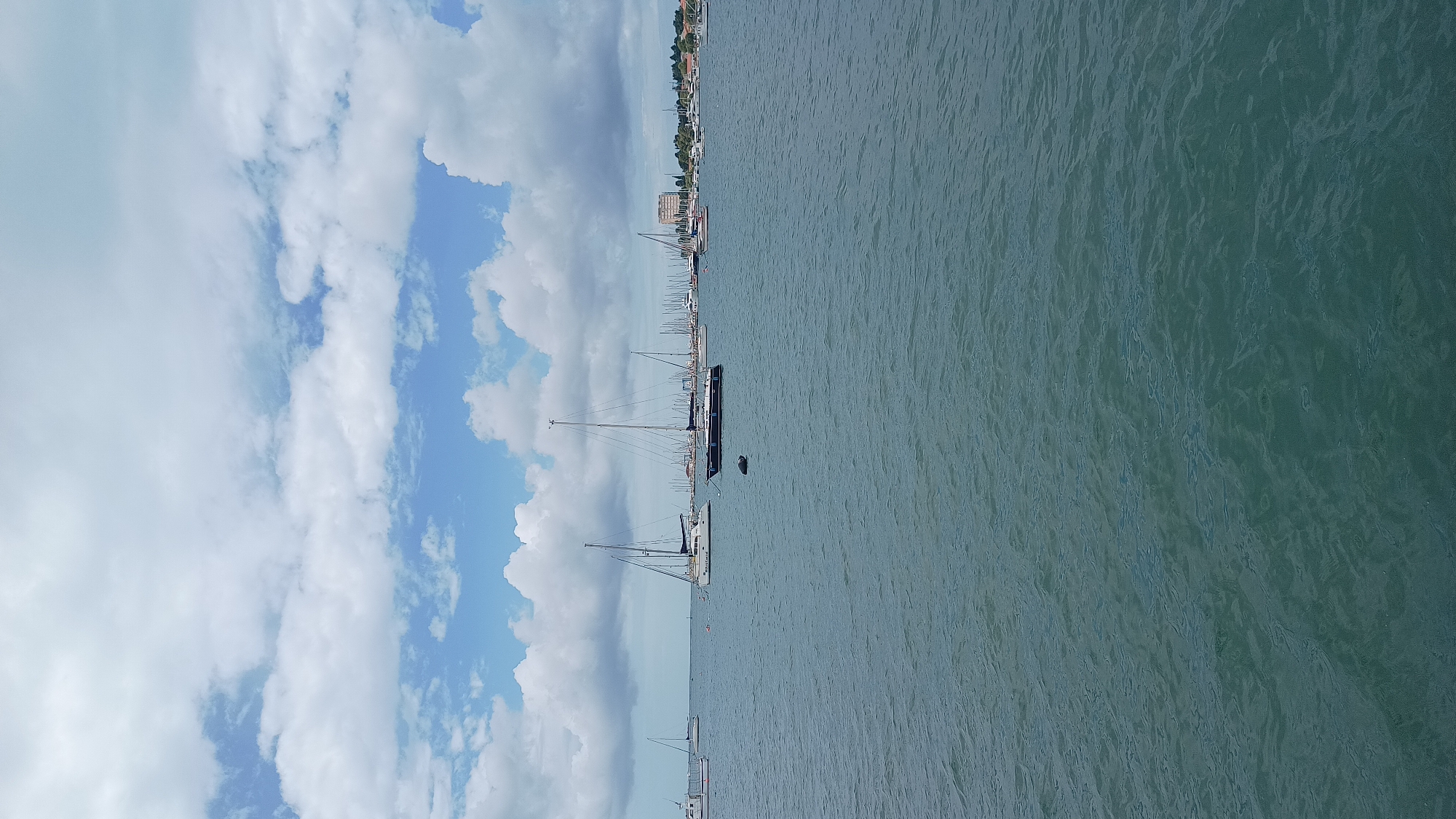 This image captures a serene maritime scene in Umag, Croatia, likely a harbor or bay. The dominant elements are the calm, deep greenish-blue sea, a partly cloudy sky, and numerous boats moored along a distant coastline.
The water surface is gently rippled, reflecting the light. Above, the sky is a mix of bright blue patches and large, fluffy white cumulus clouds, suggesting fair weather.
Many sailboats are visible, ranging in size. In the mid-ground, a dark-hulled sailboat with a tall mast is prominently featured, with a small, dark buoy or fender floating nearby. Closer to the foreground, another lighter-hulled sailboat is also visible. Further back, a long line of boats, primarily sailboats with their masts, extends along the distant shore, indicating a busy marina or anchorage.
The coastline in the background features a long stretch of land with a town or settlement, characterized by various buildings and some trees, confirming a coastal urban environment.
No people are visible in the image. The scene suggests a peaceful and quiet day, possibly late morning or early afternoon, given the bright natural light. The overall mood is tranquil and reflective of a leisurely coastal setting. There is no visible text in the image.