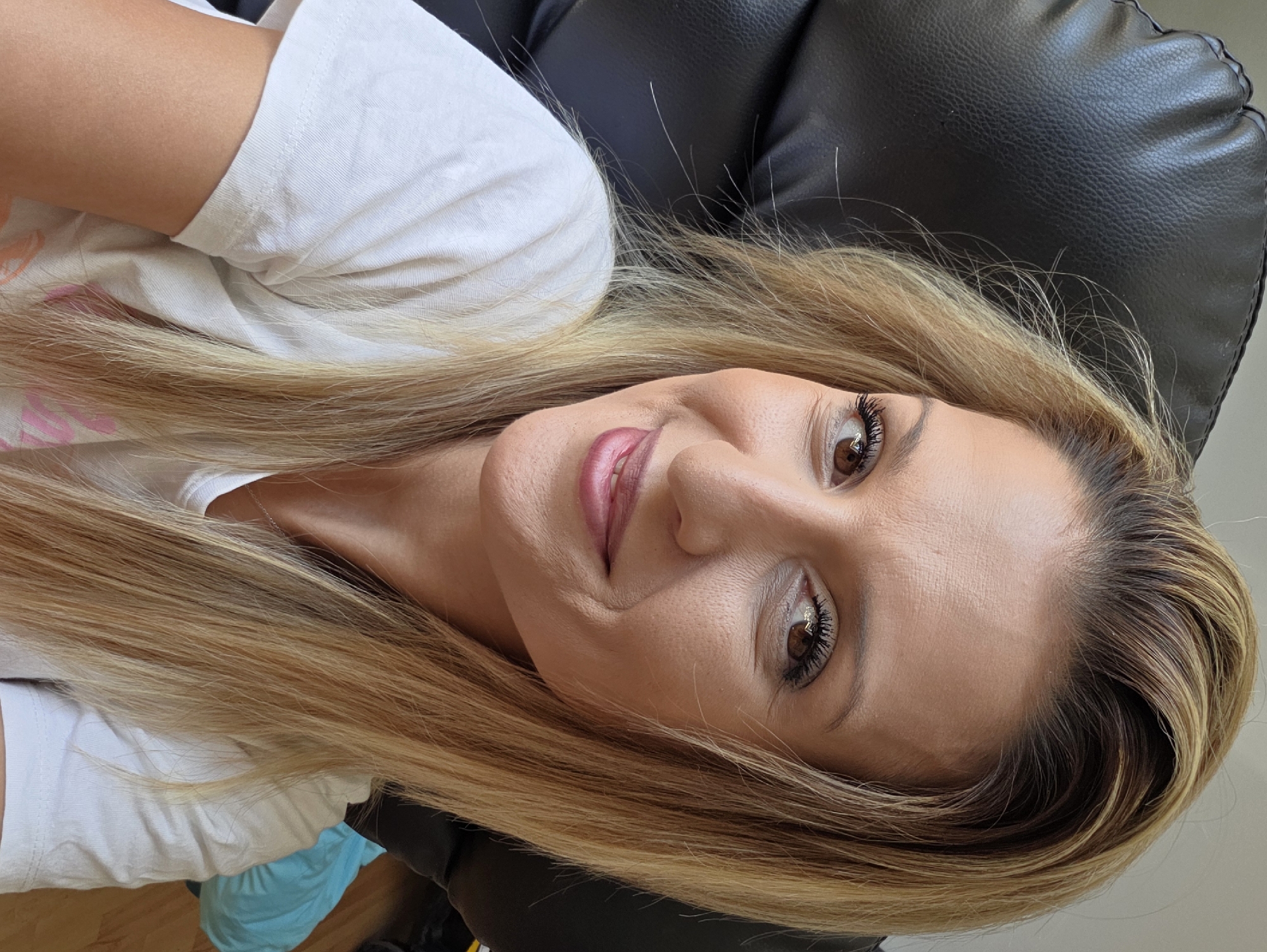 The image shows a close-up, selfie-style photograph of a woman with blonde hair, sitting in a black leather chair. She is wearing a white t-shirt. The background includes the back of the chair and a blurred floor with a light blue object. The scene appears to be indoors, likely in a residential setting. The lighting is natural and suggests daytime. There is no visible indication of the location being in Munich, Germany.