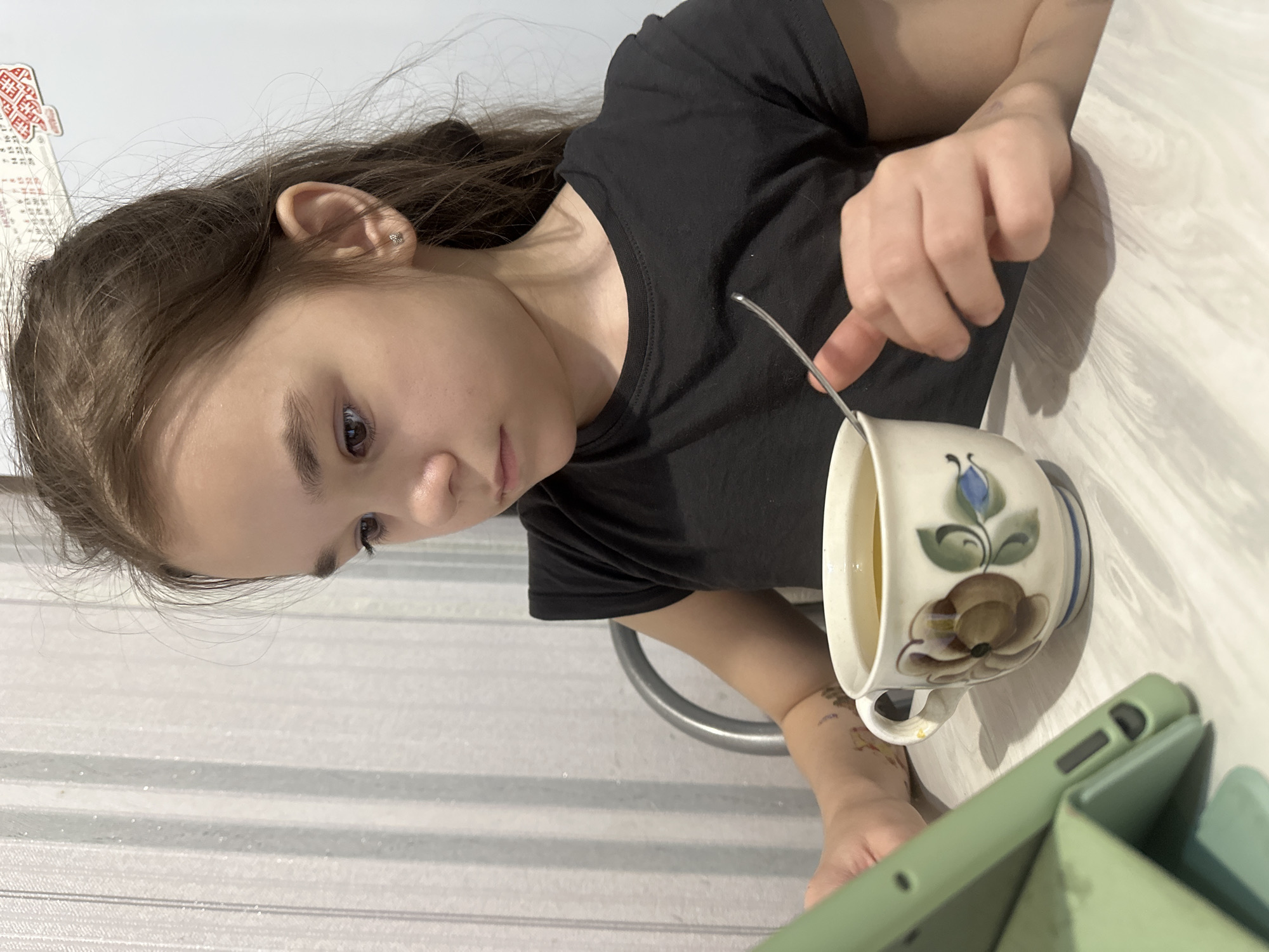 A young girl, likely a pre-teen or early teenager, with long brown hair is captured in an indoor setting, likely in Uva, Russia. She is focused on a green tablet, which is propped up in a protective case on a light wood-grain patterned table. The girl is wearing a plain black t-shirt and has a small stud earring in her visible ear. On her left forearm, a colorful temporary tattoo, possibly depicting cartoon characters or abstract shapes, is visible.
In front of her, she is stirring a beverage in a distinctive ceramic mug. The mug is cream-colored with blue and brown floral designs, specifically a blue bellflower and a brown multi-petaled flower, possibly a poppy or thistle. A metal spoon or stirrer is in the mug. Her right hand holds the stirrer, while her left hand rests near the tablet.
The background consists of a light-colored wall with subtle horizontal stripes. To the upper left, a piece of paper is taped to the wall, featuring traditional red folk art patterns and columns of text and numbers, including Cyrillic abbreviations for days of the week (e.g., "пн", "вт"), strongly suggesting it is a calendar or schedule. The lighting indicates it is daytime. The girl's expression is one of calm concentration, suggesting engagement with the content on the tablet.