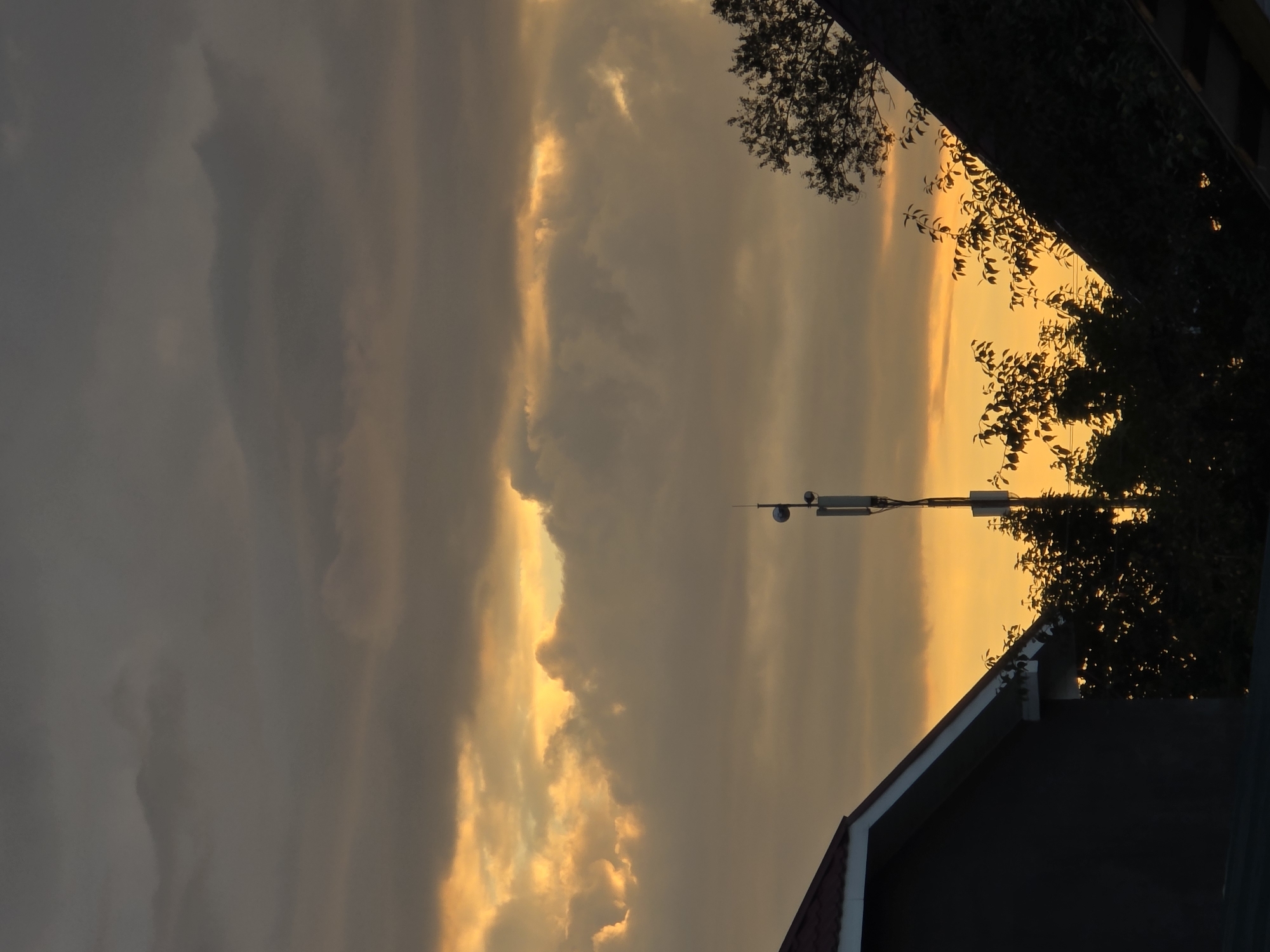 The image captures a dramatic sky at dusk, with a strip of bright orange sunlight breaking through dark, heavy clouds. Silhouetted against this vivid sky are the rooftops of two buildings, one on the left with a dark wall and red roof, and another on the right with a dark roof and lush green foliage. In the center of the frame, a slender cell tower with several antennas stands tall, also silhouetted. The surrounding trees are dark shapes, their leaves barely discernible. The overall impression is one of an approaching evening in a somewhat urban or suburban setting, with the intensity of the sunset dominating the scene. There is no indication of the city of Речицы, Russia within the image itself.