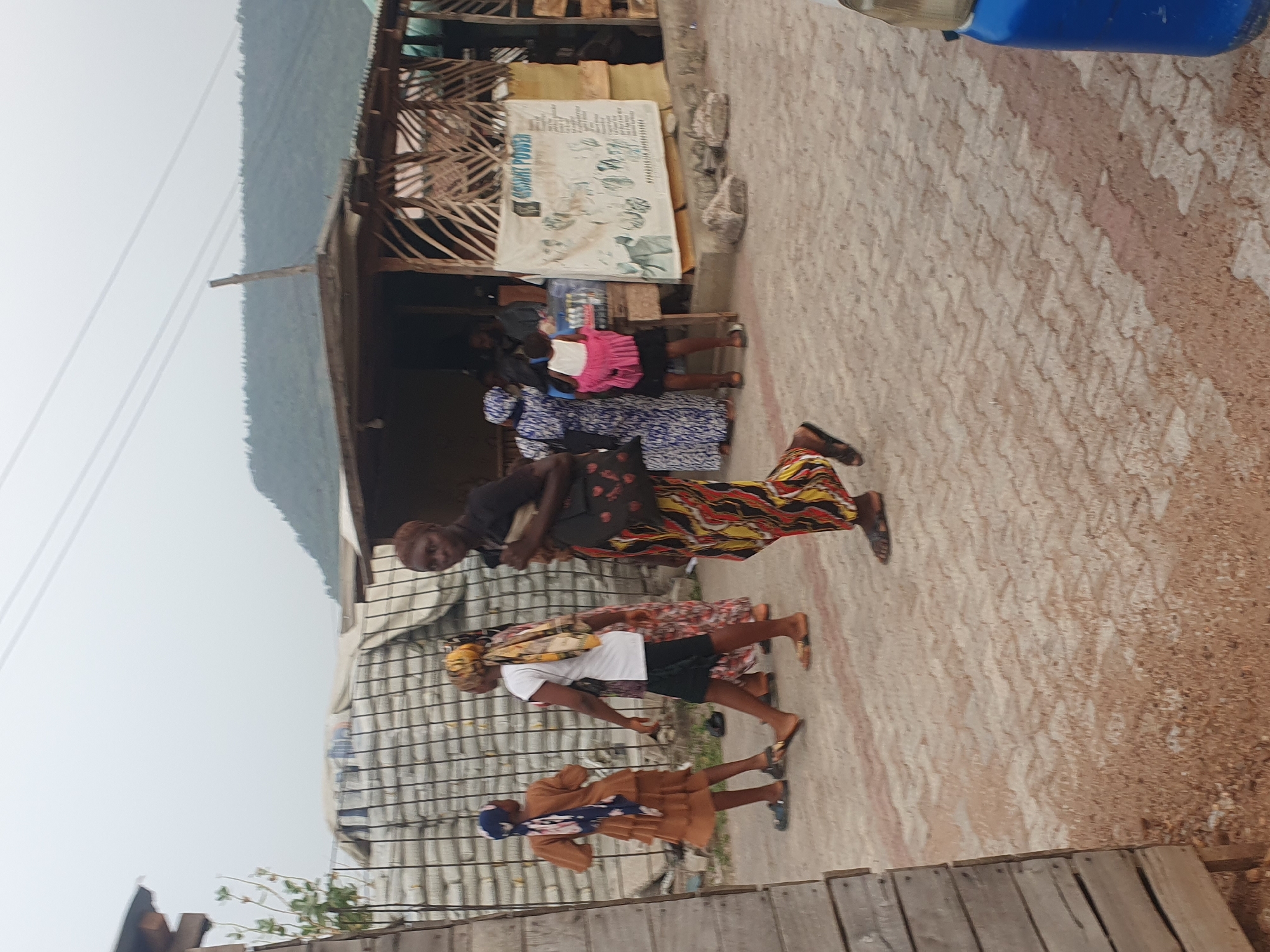 The image captures a vibrant street scene in what appears to be a semi-urban or rural area of Lokoja, Nigeria, under daylight conditions. The sky is bright, suggesting either clear skies or light overcast.
The primary subjects are several individuals, mostly women, walking along a paved path that transitions into a dirt road. In the foreground, a woman wearing a black top and a long, colorful patterned skirt (featuring yellow, red, and black) walks towards the left, wearing sandals. Behind her, another person, also in sandals, wears a white t-shirt, dark shorts, and a patterned headscarf, walking in the same direction. Further back, a woman in a brown dress and a blue head covering is seen walking. To the far left, near a building, a child in a pink top and dark shorts stands, alongside a person in a blue and white patterned dress.
The setting includes simple, single-story structures lining the path on the left. These buildings feature wooden frameworks, some with decorative lattice work, and possibly corrugated metal roofing. A large white sign is prominently displayed on one of these buildings, bearing the legible text "OSMIT POWER" and various illustrations of body parts and abstract designs, suggesting it might advertise health products or services. In the distance, undulating hills or mountains are visible under the bright sky, with faint power lines traversing the scene. The ground transitions from a light-colored, patterned paving to a reddish-brown dirt track, indicative of the local environment.