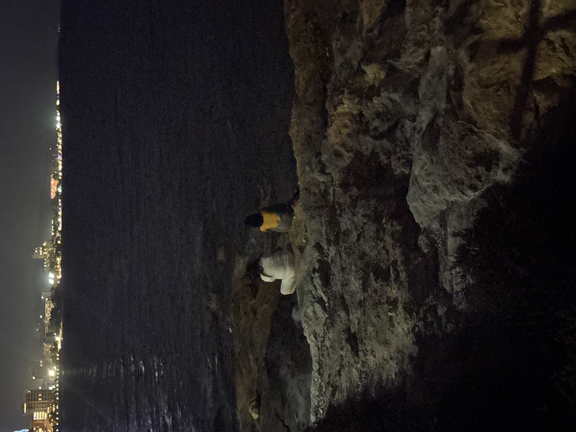 The image depicts a nighttime scene in the Agios Tychona Community, Cyprus. In the foreground, two individuals are seated on rocks near the water's edge. One person, wearing white attire, is positioned to the left, while another, in a yellow shirt, sits to the right. Beyond them, the dark water stretches out, reflecting distant city lights. In the background, the Agios Tychona coastline is illuminated by numerous buildings and lights, suggesting a developed urban area. The overall atmosphere is tranquil and dark, with the city lights providing a point of visual interest against the night sky and water.