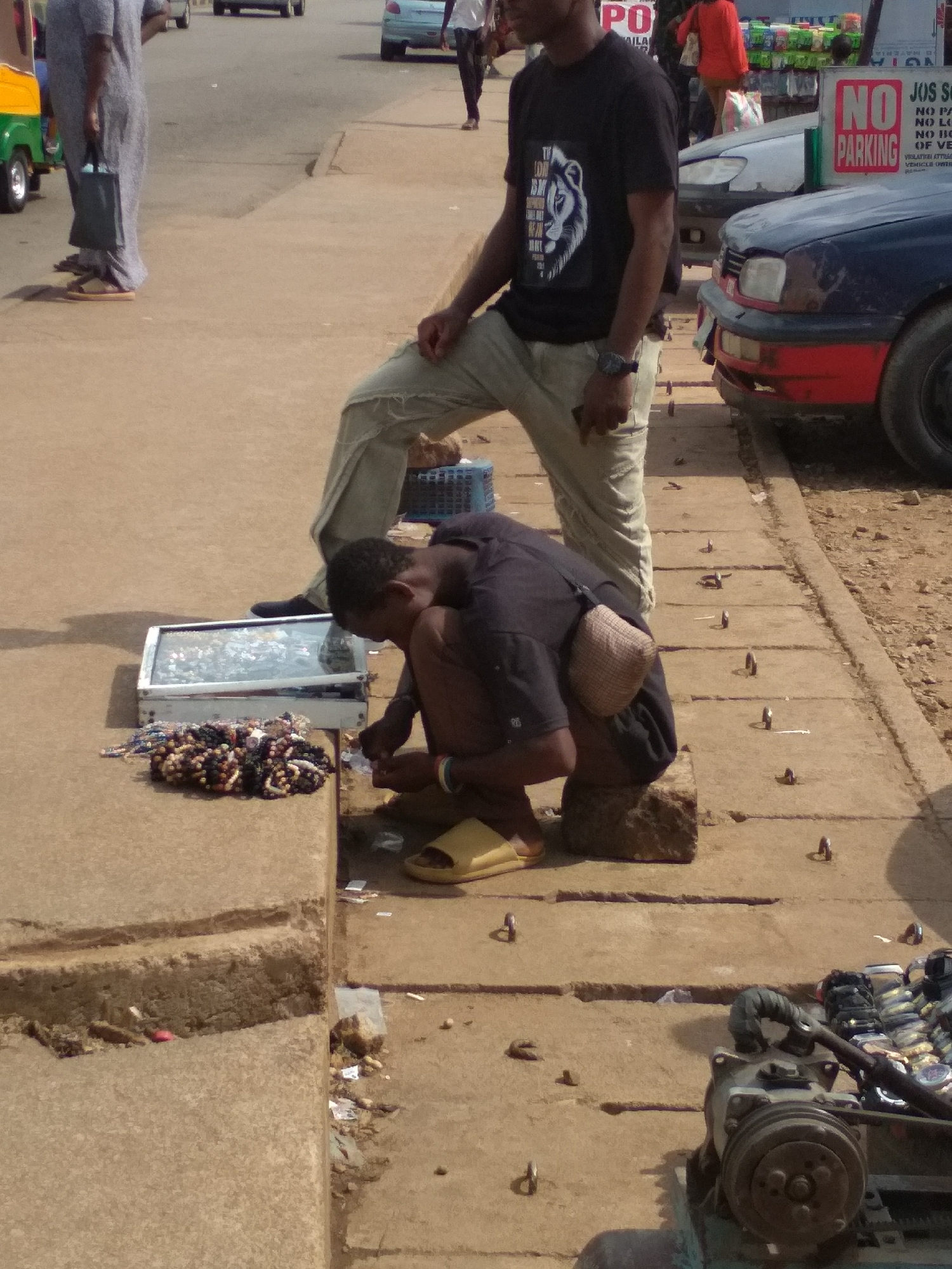 This image captures a daytime street scene in Barakin Lerawi, Nigeria, under bright, sunny conditions. The foreground features three men on a concrete sidewalk.
On the far left, a man dressed in a light-colored, long traditional garment and sandals walks past, carrying a dark bag. In the center, another man stands with his right foot elevated on a lower ledge. He is wearing a black t-shirt featuring a white lion graphic and partial text that includes "TI LON SM PO", light-colored ripped jeans, and black shoes, with a watch on his left wrist.
To the right of the standing man, a third man is crouched on a small stone, attending to items displayed on the ground. He wears a dark t-shirt and light slippers, with a small, light-colored cross-body bag on his back. In front of him are a display case with various items and a pile of colorful beaded jewelry, likely bracelets or necklaces, suggesting he is a street vendor.
The background reveals a paved road with traffic, including a green and yellow tricycle (keke napep) on the left and two cars on the right—one light-colored and one dark blue with a red bumper. Pedestrians are also visible in the distance.
A prominent red and white "NO PARKING" sign is on the right side of the road, alongside another sign with fragmented text reading "JOS SO", "NO PA", "NO LO", "NO BO", "OF VE", and "VEHICLE OWN". The pavement on the right features a series of small metal rings or hooks embedded in the concrete, and a piece of machinery is visible in the bottom right corner. The scene depicts a typical moment of urban activity and street commerce.