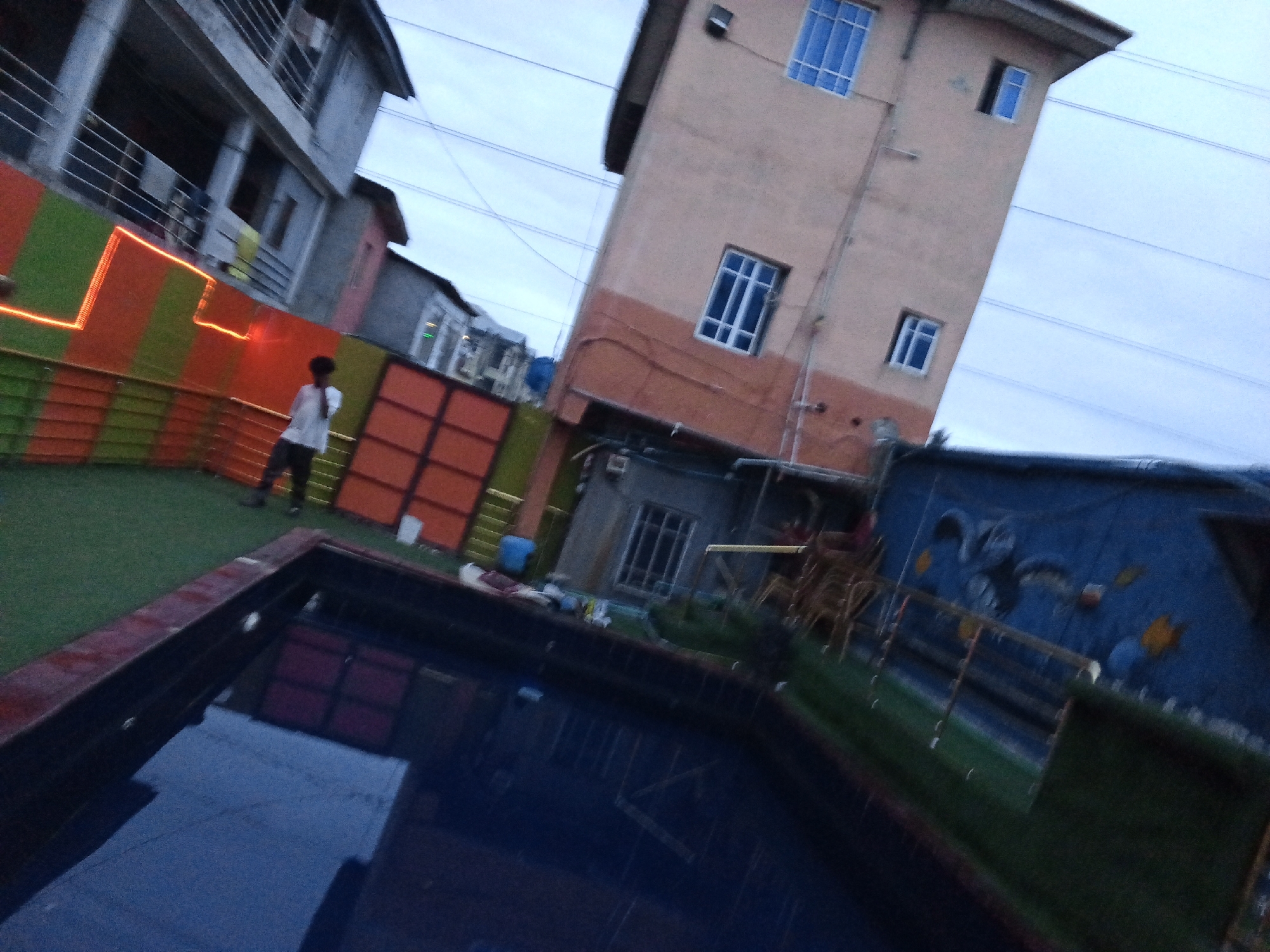 The image shows a courtyard area with a swimming pool in the foreground, an artificial turf lawn, and several buildings in the background. A person wearing a white t-shirt and dark pants stands on the artificial turf near a multi-colored fence. The fence is composed of vertical orange and green panels with horizontal orange lighting accents. Behind the fence, a multi-story building with balconies is visible. To the right, a taller, light-pink building has several windows. A blue mural with yellow accents is painted on a structure on the far right. The scene appears to be at dusk, with the sky overcast. The swimming pool is filled with dark water and reflects the surrounding environment. The location is identified as Ado, Nigeria.