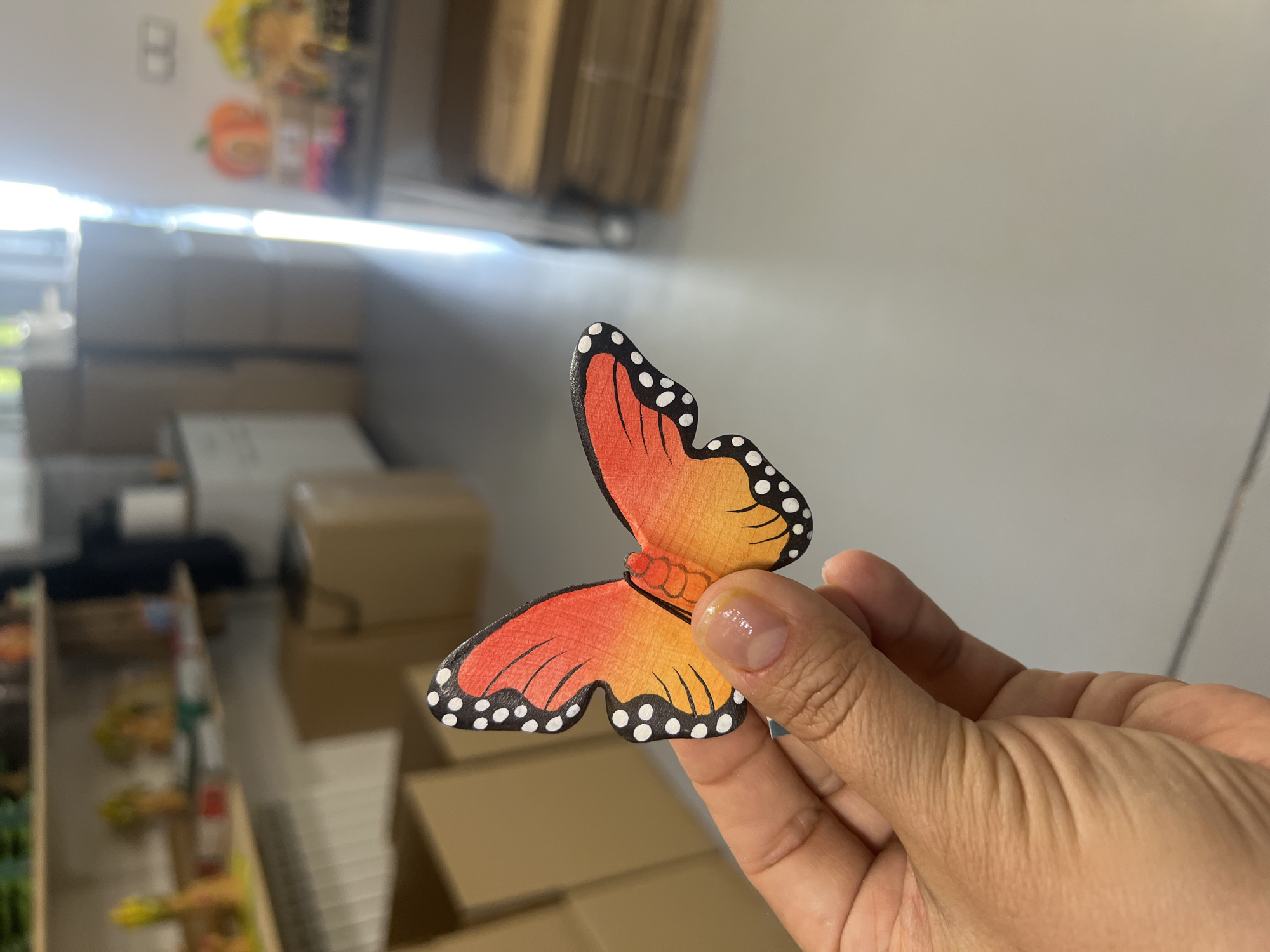 A person's hand holds a wooden butterfly decoration. The butterfly is painted with orange and yellow gradients and has black and white dots along its wings. The background appears to be an indoor space with shelves, boxes, and a bright light source coming from the right. The location is a room in an unknown city and country.