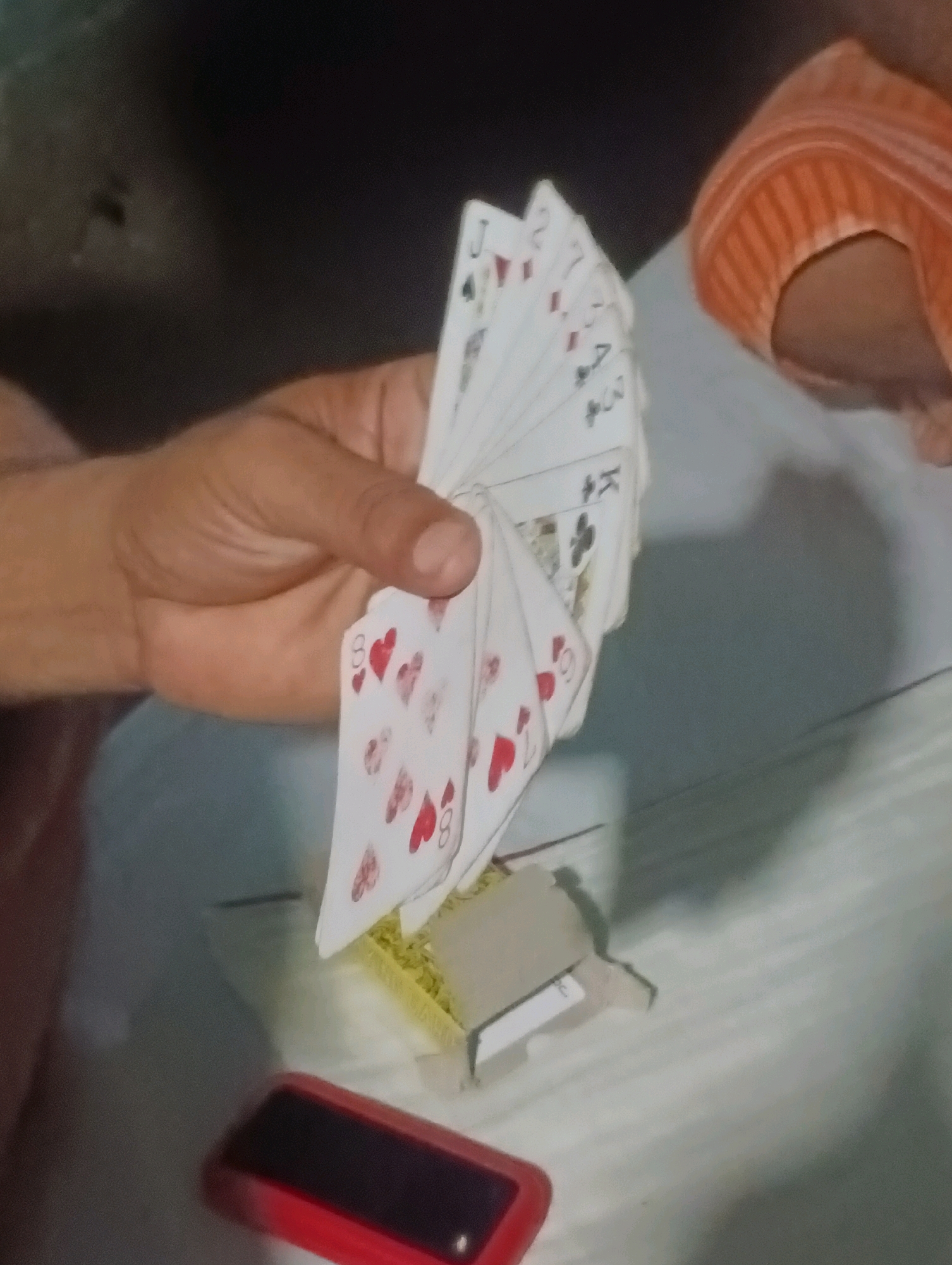 The image shows a close-up of a person's hand fanning out a deck of playing cards. The cards are white with red and black suits and numbers. Some of the visible cards include a Jack of Spades, a 2 of Spades, a 7 of Hearts, and an 8 of Hearts. The hand holding the cards is light-skinned with visible knuckles and finger creases. To the right of the hand, a piece of orange fabric, possibly a towel or blanket, is draped over an unseen surface. In the foreground, a red object with a black rectangle in the center is visible, likely a mobile phone. The background is dark and out of focus. The scene appears to be indoors, with a light-colored, textured surface beneath the objects, possibly a table or bed. There is no specific indication of the city of Kesari, India, in the image.