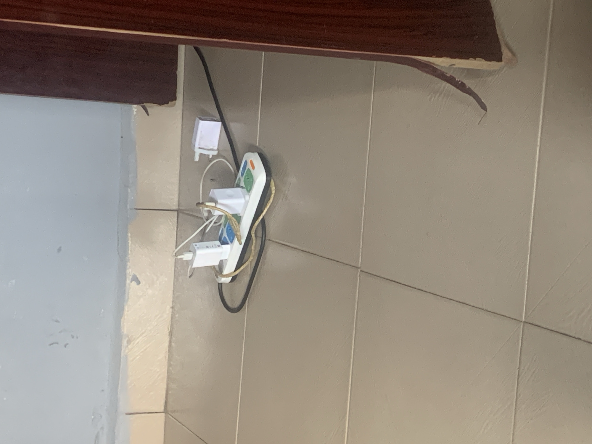 The image shows a close-up view of a white power strip with multiple electrical adapters plugged into it. The power strip is mounted on a tiled wall, with beige tiles forming the background. Above the wall, a dark brown wooden surface is visible, possibly a shelf or a cabinet.
Several white electrical adapters are plugged into the power strip. One of them appears to be a USB charger with multiple ports and some colored indicator lights. Other adapters are of different shapes and sizes. A black electrical cord is connected to the power strip and runs upwards, disappearing out of frame. Another white adapter, with a plug on one side, is plugged into a wall socket above and to the left of the power strip. This adapter is also connected to a white wire.
The setting appears to be indoors, likely in a residential or commercial space. The lighting suggests it is daytime, but there are no strong shadows that would indicate a specific time of day. The image is taken at an angle, making it difficult to determine the exact orientation of the room. The overall impression is one of a functional setup for charging electronic devices.
