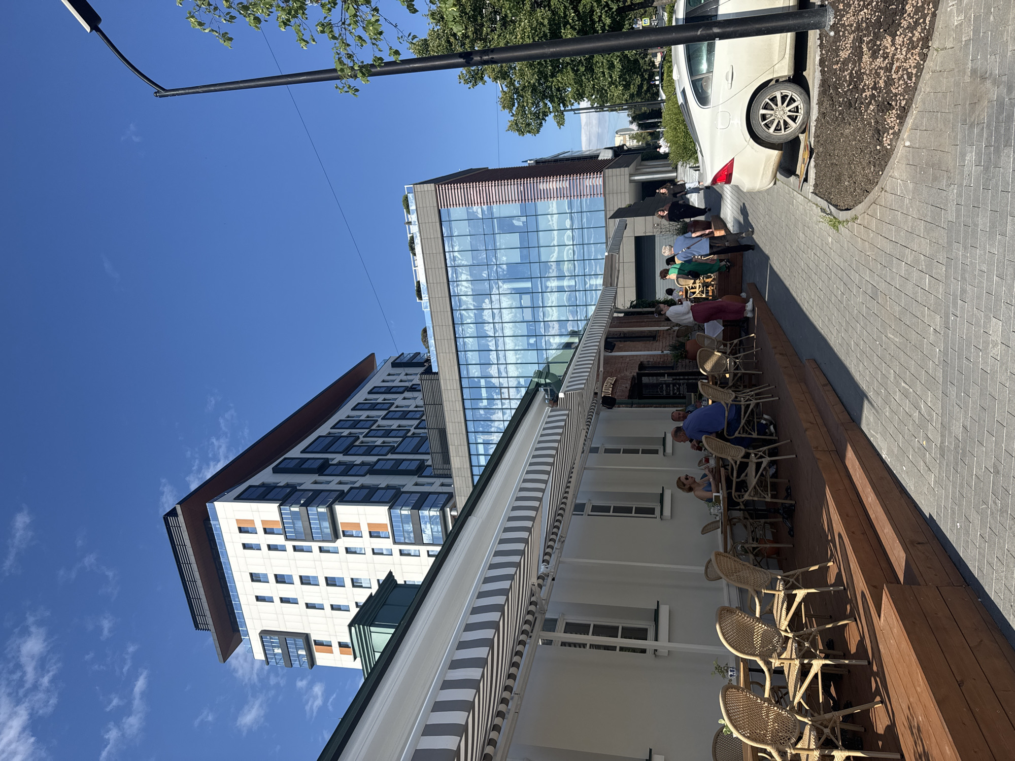 In Yekaterinburg, Russia, this image depicts an outdoor seating area of a cafe or restaurant. Several people are visible, seated at tables and walking along the sidewalk. The scene is set on a sunny day, with a clear blue sky and some light clouds.
The foreground features a wooden deck with numerous wicker chairs and tables, suggesting a leisurely atmosphere for dining or socializing. A striped awning provides shade over part of the seating area. To the left, a modern building with a glass facade reflects the sky and surrounding environment. Further left, another contemporary building with many windows rises, its architecture contrasting with the lower, more traditional-looking structure in the immediate foreground.
In the background, a white car is parked alongside the sidewalk, which is paved with cobblestones. Green trees line the street, adding to the urban scenery. The time of day appears to be daytime, likely late morning or early afternoon, given the bright sunlight casting shadows. The overall mood seems relaxed and pleasant, indicative of a typical day in a city with outdoor dining options. No visible text is readily discernible in the image.
