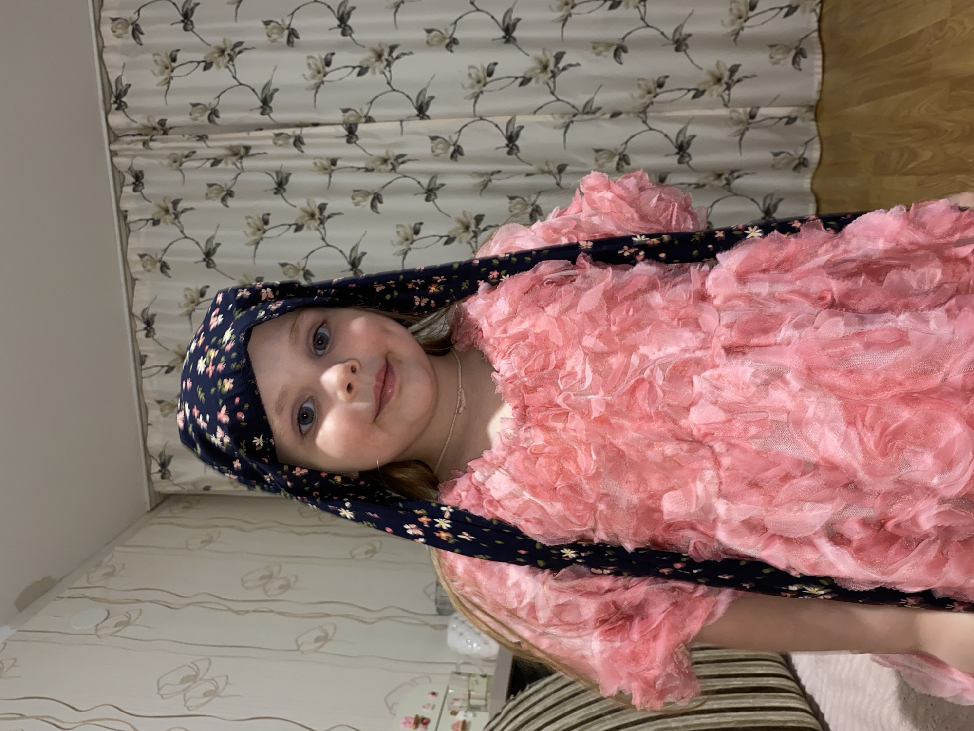 An image, presented sideways but depicting a scene in a vertical orientation when corrected, features a young girl with light blonde hair and blue eyes. She is looking directly at the camera with a gentle, engaging smile. The girl is wearing a dark blue headscarf adorned with a pattern of small, multicolored flowers. Her main attire is a voluminous, vibrant pink dress characterized by multiple layers and intricate ruffled fabric details, giving it a rich, textured appearance.
The setting is an indoor space, likely a room within a home in Votkinsk, Russia. Behind and to the left of the girl are curtains with a light background, featuring a repeating pattern of dark, stylized floral branches and leaves. To her right, a section of light-colored wooden flooring is visible. Below the wooden floor and extending to her lower right is wallpaper with a beige background, patterned with subtle wavy brown lines and small, oval-like designs. In the bottom left corner, a white piece of furniture, possibly a dresser or shelf, is partially visible, topped with small, decorative items. A striped textile, appearing to be a cushion or part of bedding, occupies the bottom right corner of the frame. The scene is evenly and brightly lit, suggesting a well-lit interior. The girl's expression conveys a sense of calm and happiness. No visible text is present in the image.