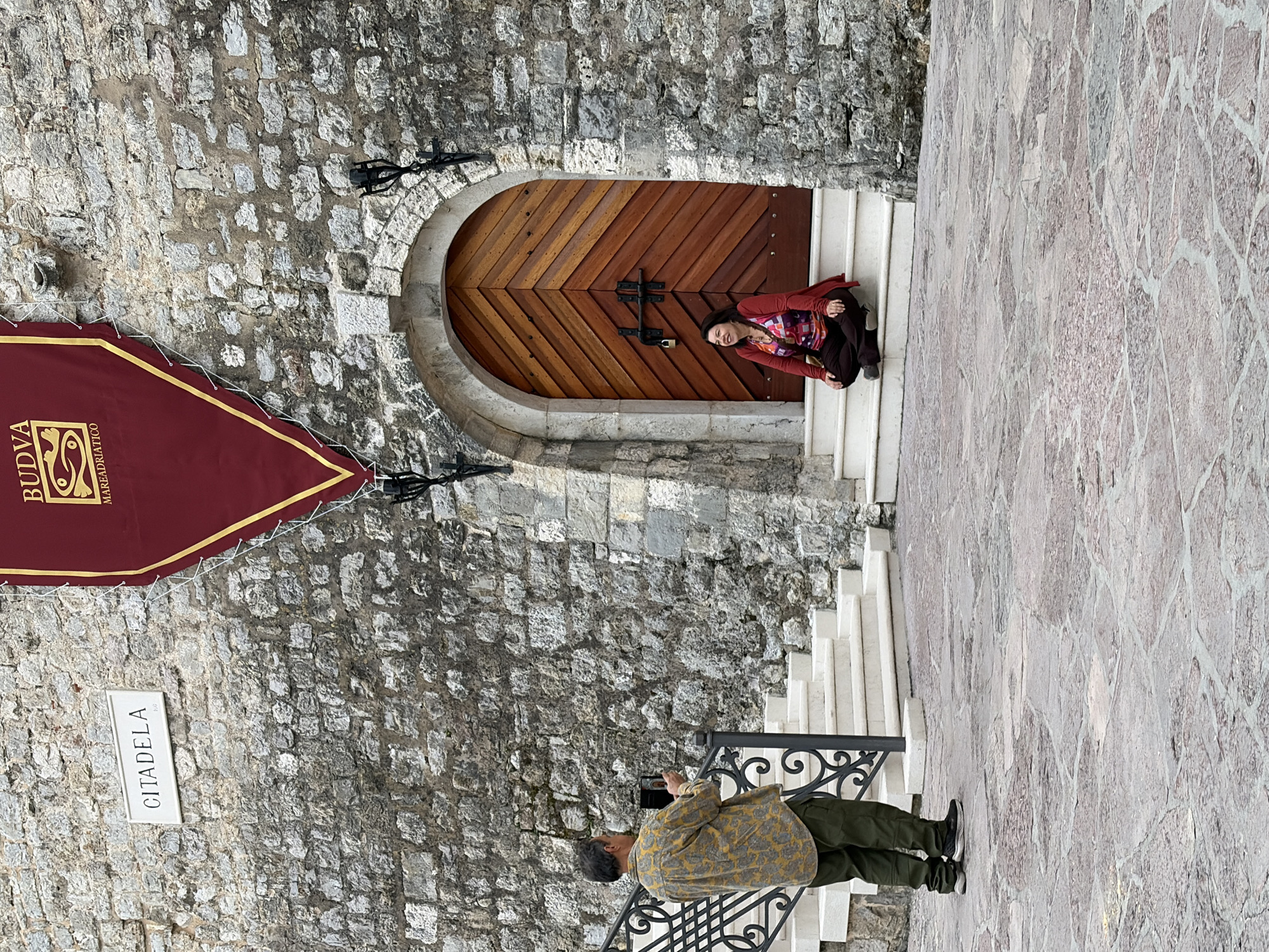 A middle-aged woman is kneeling on the steps in front of a large, arched wooden door of a stone structure, appearing to pose for a photograph. She is wearing a reddish-brown jacket, a patterned top, and dark pants. A man stands below her on a landing, facing her, holding a camera, apparently taking a photo. He is wearing a patterned, light-colored shirt and green pants. The stone building features a red banner with "Budva" and "Mare Adriatico" printed on it. A sign reading "Citadela" is visible on the stone wall. The location is in Budva, Montenegro, as indicated by the banner. The environment consists of stone structures, likely part of the old city walls or a historical site, and the cobblestone ground.