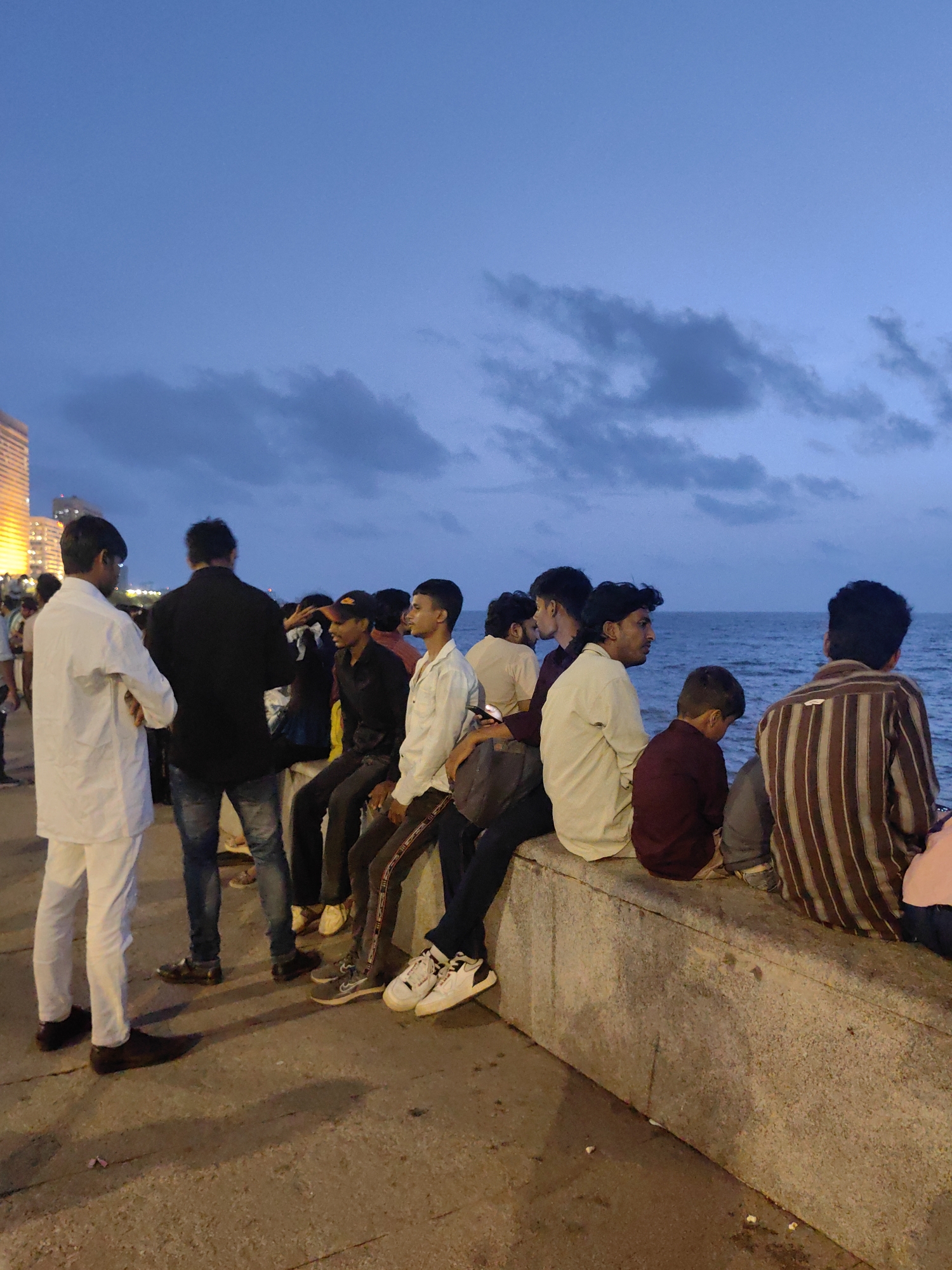 An outdoor, medium shot captures a group of individuals gathered along a concrete seawall by the ocean at dusk in Mumbai, India. The sky above is a deep blue with some scattered clouds. On the left, two men stand facing each other, one in a white outfit and the other in black. The majority of the group is seated on the seawall, facing the ocean. Their attire consists of casual clothing, including shirts, pants, and sneakers. The ocean, visible in the background, appears calm. To the far left, a tall building with illuminated windows is partially visible, suggesting an urban setting within Mumbai. The ground in front of the seawall is paved and appears damp in places, with some small debris scattered about. The overall atmosphere suggests a relaxed evening gathering by the sea.