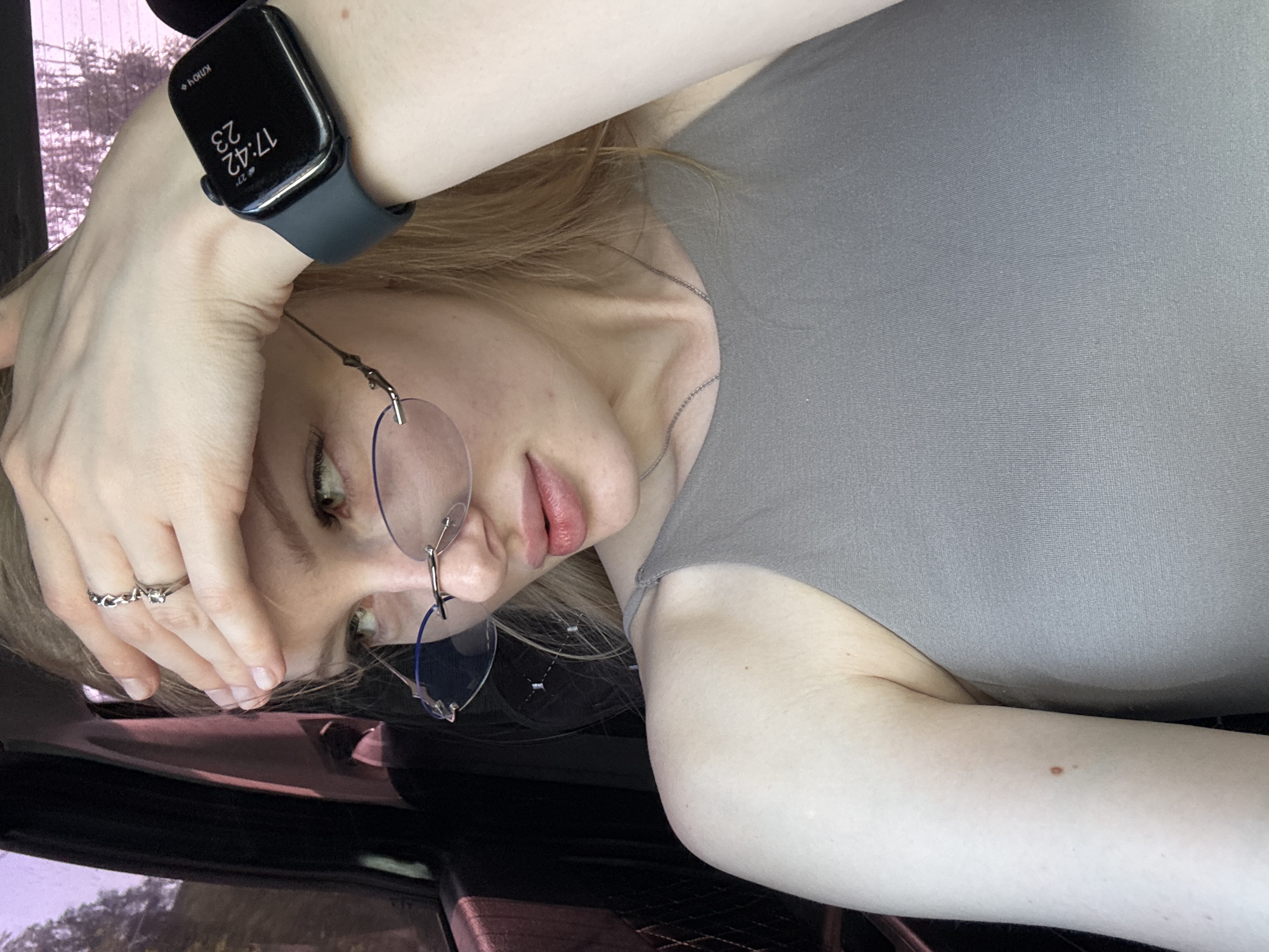 A young adult female with fair skin and blonde hair is depicted from a high-angle perspective. The subject is partially visible, and tilted to the side. She is wearing a gray shirt. The woman is wearing glasses. A digital wristwatch is visible on her left wrist, displaying the time as 17:42. She is wearing a silver ring. The environment appears to be an interior, possibly a car. The location, Perm, Russia, is not discernable from the available visual information.