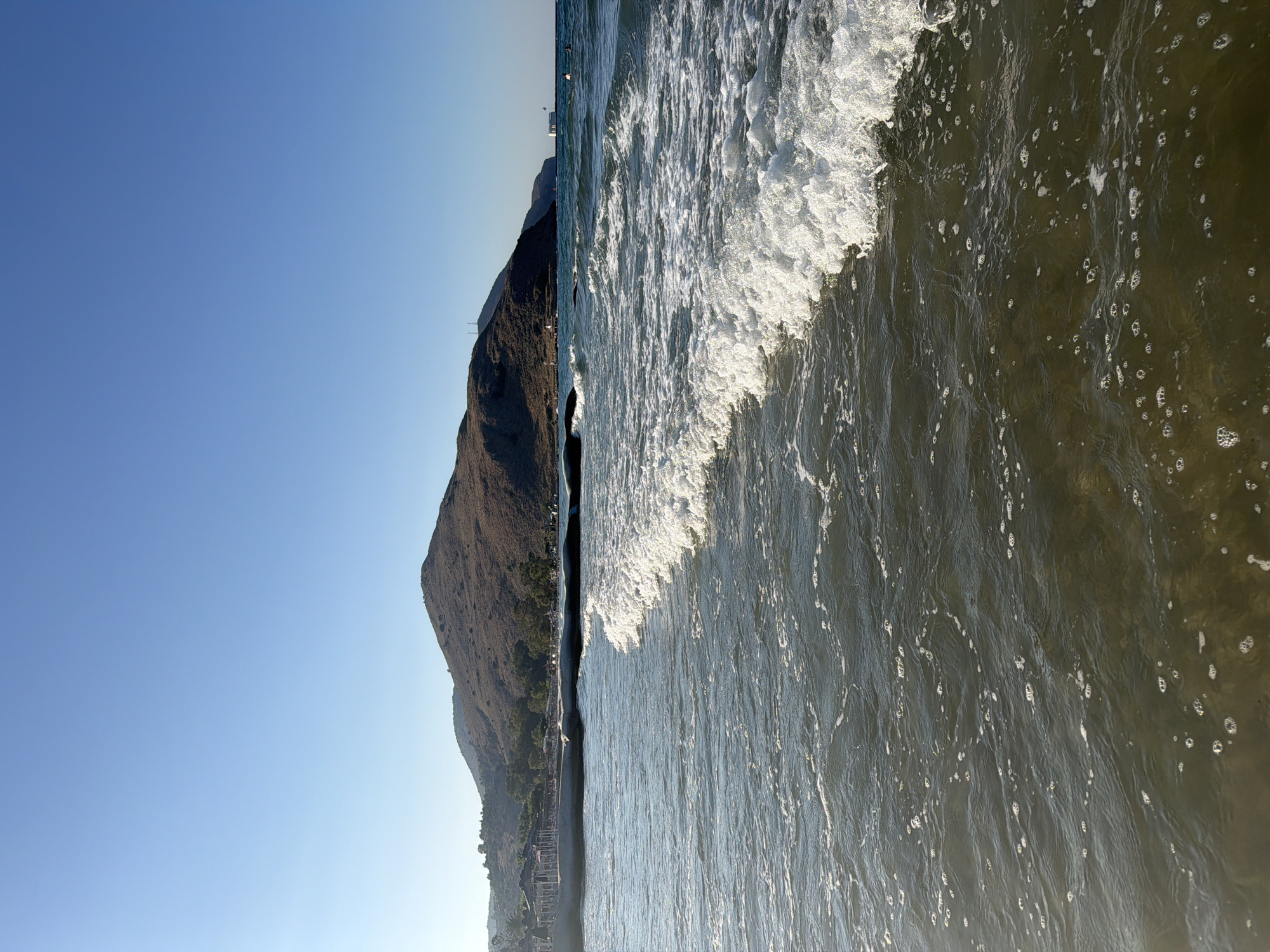 The image depicts a serene beach scene in Georgioupoli, Greece, under a clear blue sky. In the foreground, gentle waves with frothy white crests roll onto a shallow, somewhat murky shore, revealing scattered bubbles on the water's surface. A dark, submerged structure, possibly a breakwater, is partially visible near the left side of the frame.
Further inland, a paved walkway or pier is lined with thatched umbrellas, suggesting a developed beach area with amenities for visitors. Beyond the walkway, a cluster of trees adds a touch of greenery to the landscape. The background is dominated by a large, arid hill covered in dry vegetation, characteristic of the Mediterranean terrain. In the distance, a white building with a red roof stands near the water's edge, and a few other distant structures are visible on the coastline. Two individuals are present in the water to the right, appearing to swim or float. The overall impression is one of a tranquil day at the seaside, with a clear demarcation between the tranquil sea and the dry, hilly landscape.