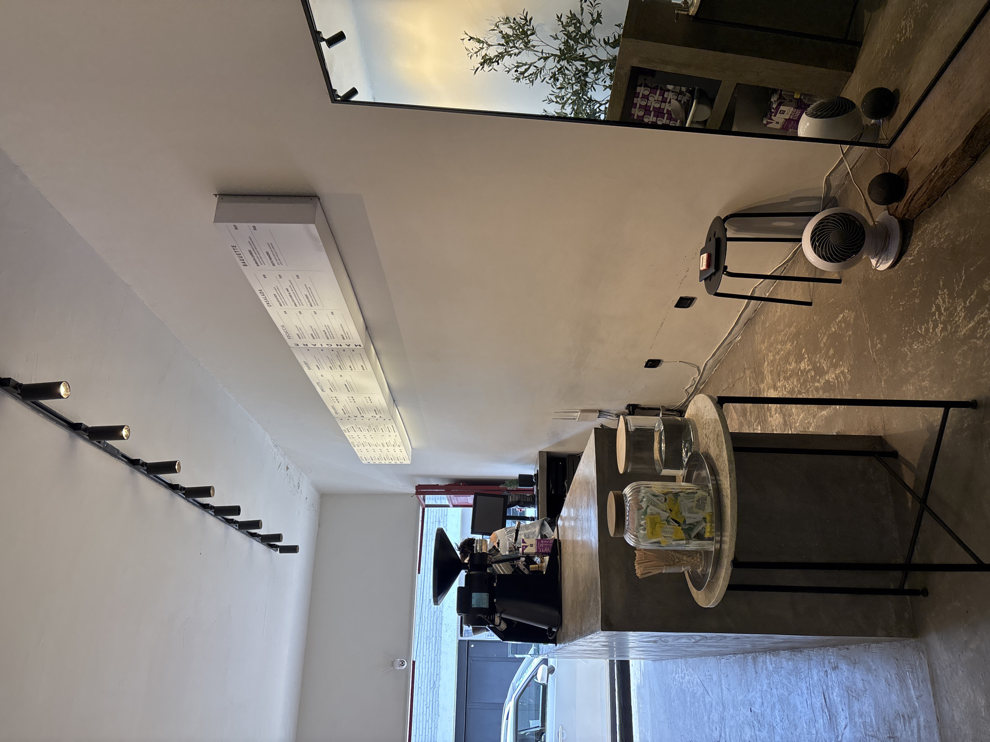 The image depicts the interior of a business, possibly a cafe or coffee shop, located in San Andrés Cholula, Mexico. The scene is shot indoors.
On the left wall is a track lighting system with several cylindrical lights mounted along it. Adjacent to the lights, a menu board is affixed to the wall, listing various food and drink options.
In the center of the image, a counter is visible, equipped with a coffee machine. Behind the counter is a view into what appears to be the outside, revealing a vehicle and elements of the urban landscape.
To the right, a round table with black metal legs stands. The table has glass containers or jars of sugar packets on top. A mirror is mounted on the right wall, reflecting the outdoor scenery. Two small, white fans are also on the wall by the mirror.
The overall impression is a contemporary business in an urban environment.