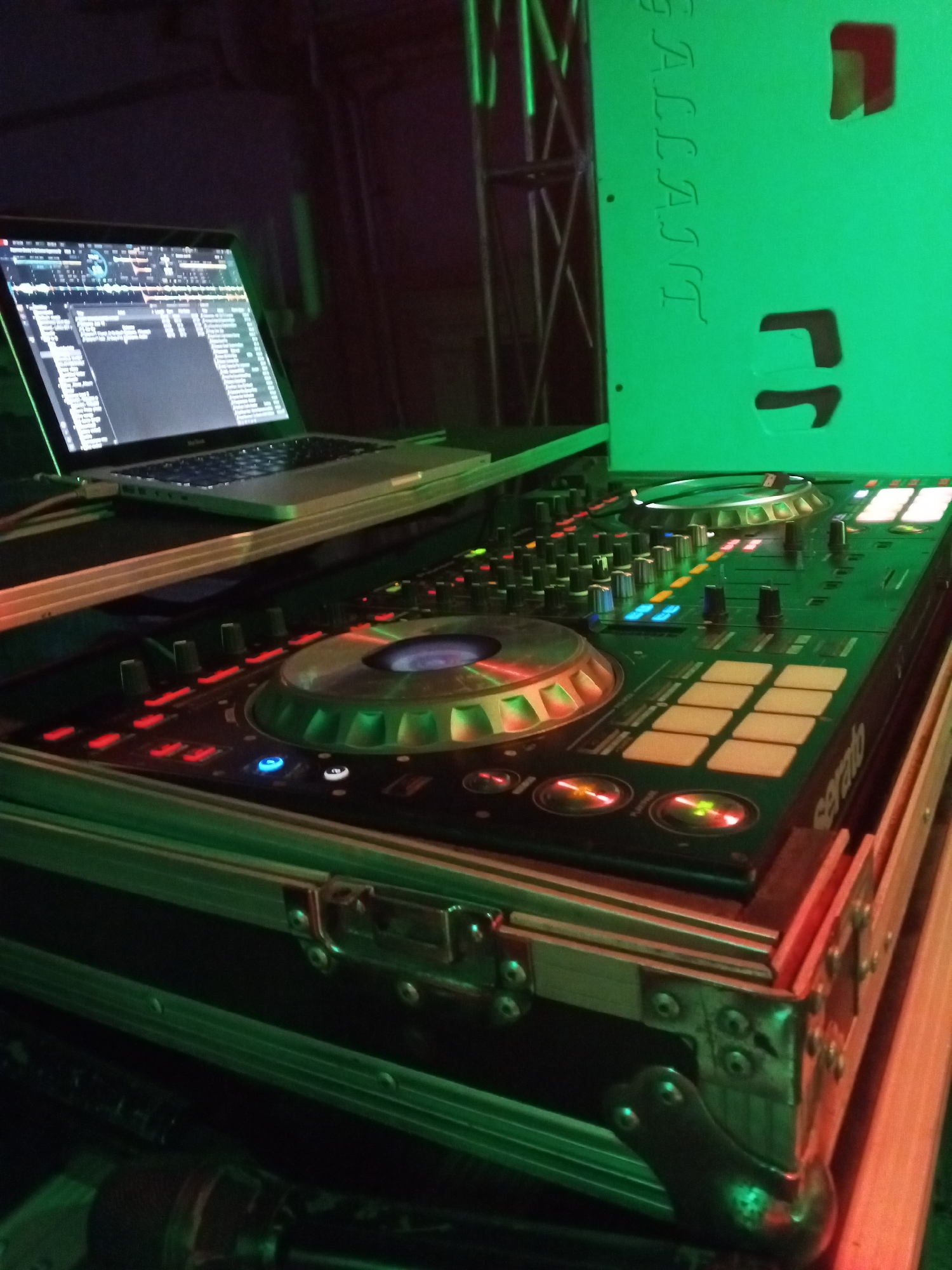 The image depicts a DJ setup indoors. A laptop sits on a shelf above a DJ controller. The DJ controller has multiple buttons, knobs, and jog wheels, with the brand name "serato" visible. The controller is enclosed in a road case with metal corners. To the right of the setup is a vertical structure with the word "GALLANT" displayed on it. The room appears to be dimly lit, with a green hue cast across the equipment. The image does not contain any identifiable information to verify if it is located in Lokoja, Nigeria. No people are visible.