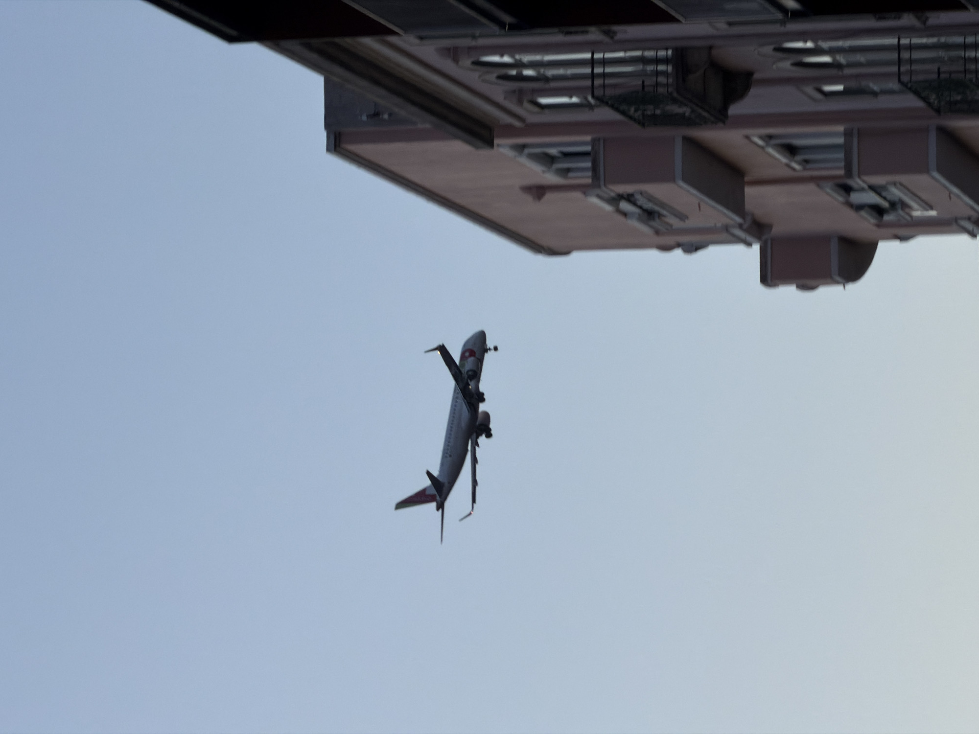 The image shows an airplane flying close to a building. The plane is a passenger jet, likely descending for landing with its landing gear deployed. The building appears to be multi-story with balconies and detailed architectural elements. The color palette is dominated by grays and off-whites, set against a light blue sky. Given the proximity of the building to the path of the aircraft and based on visual characteristics, the image likely depicts a scene near Lisbon Airport (LIS) in Portugal, where low-flying planes are commonly seen near urban areas during approach and departure. The buildings, the positioning of the plane close to the buildings and the general angle and positioning of the photo support this conclusion.