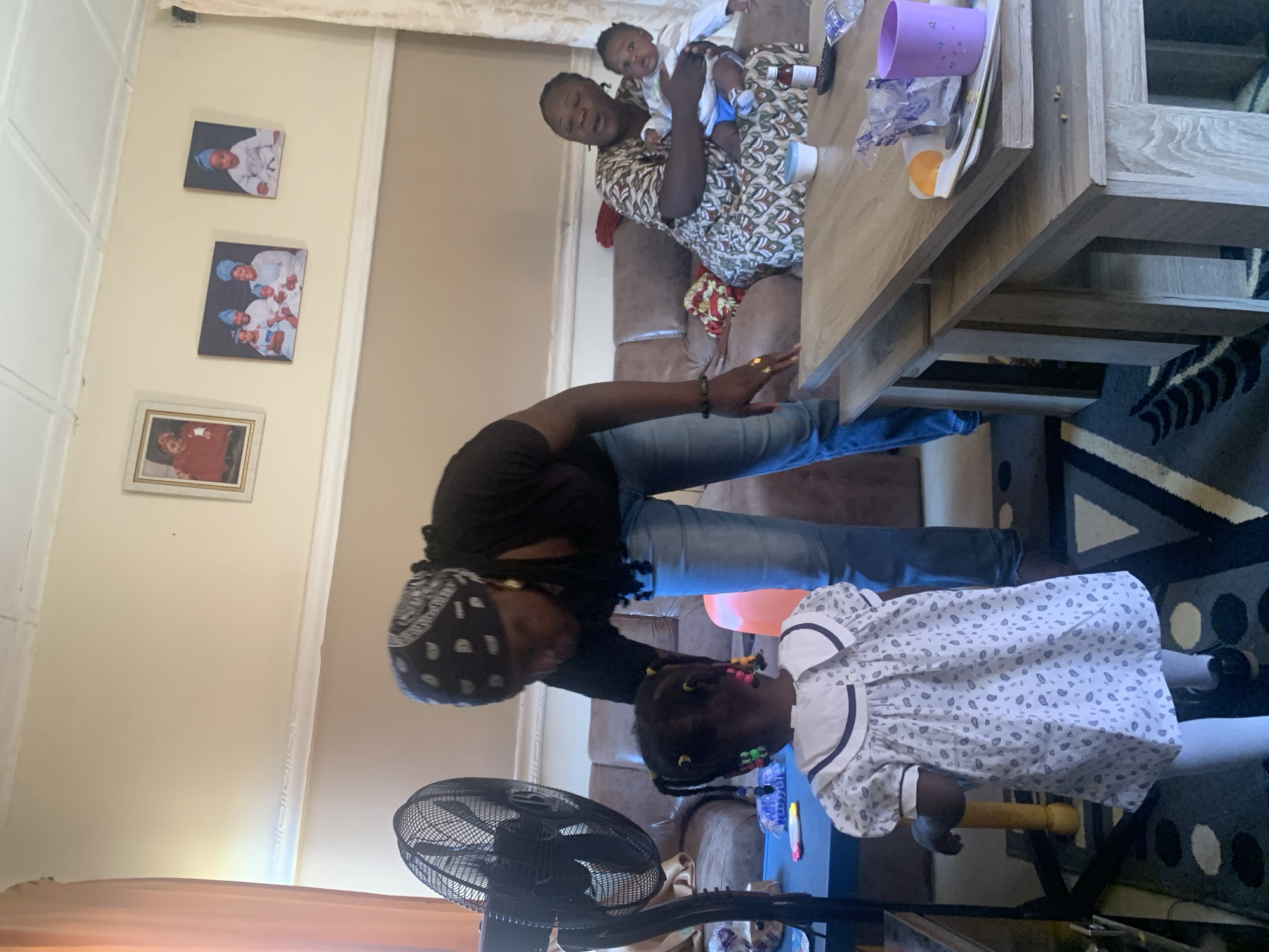 In Gura Top, Nigeria, an indoor scene depicts a living room with a family. A woman wearing a black bandana and black top, along with blue jeans, is bent over interacting with a young child in a white dress with a patterned print. The child has braided hair with colorful beads. To the right, another woman sits on a brown couch, holding a baby. The room is furnished with a couch, two coffee tables, and a standing fan. Framed pictures of a family are displayed on the wall. The overall impression is a casual family gathering.