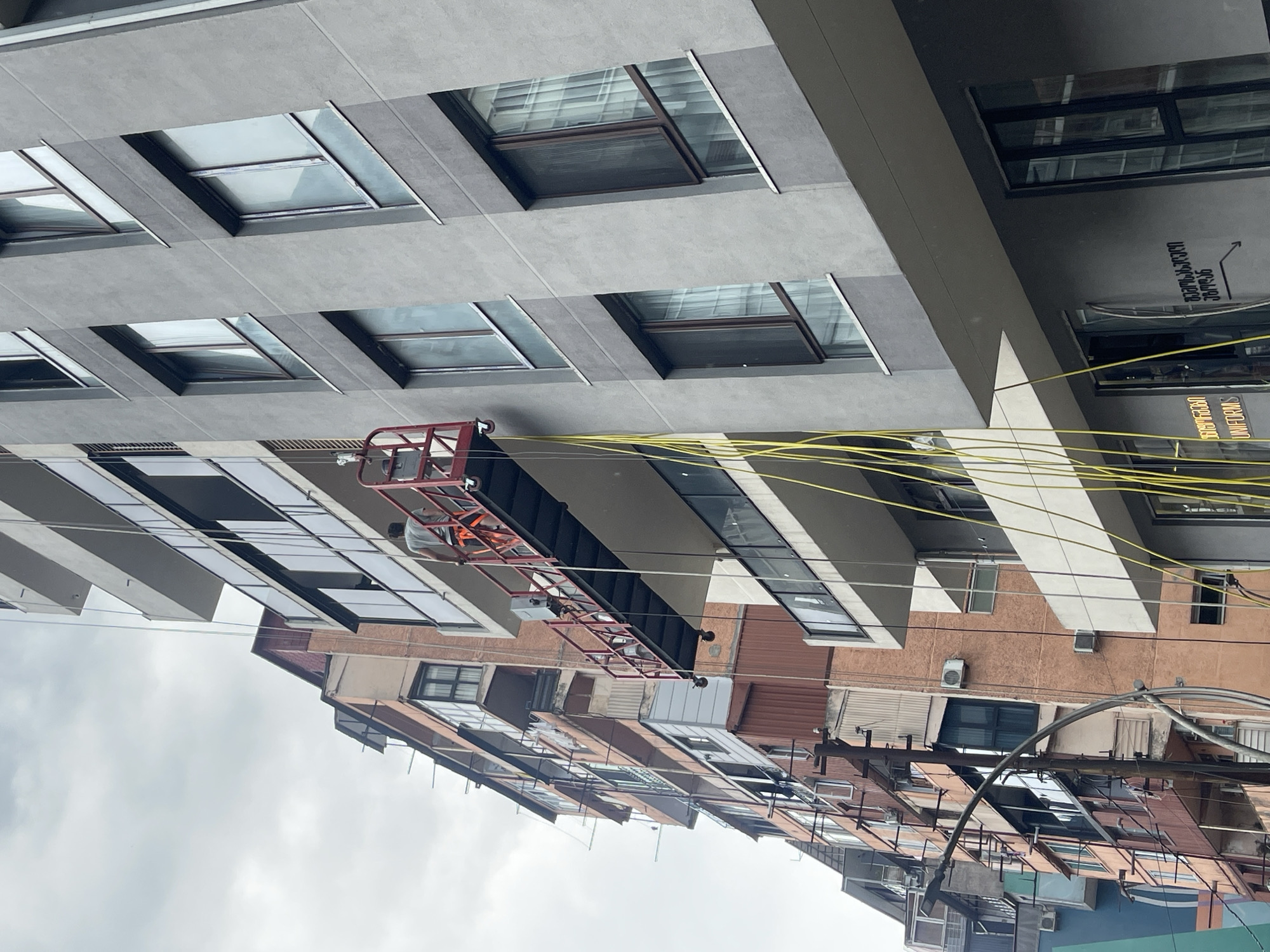 The image captures a street view in Batumi, Georgia, featuring a scaffolding platform suspended by yellow cables against the backdrop of two buildings. One building is a modern, grey-faced structure with multiple windows, while the adjacent building is older, with a more textured, pinkish-orange facade and numerous balconies. On the scaffolding, a construction worker, dressed in a grey t-shirt and orange safety harness, is visible, actively engaged in work. The sky is overcast, contributing to the diffused lighting of the scene. The presence of power lines and streetlights suggests an urban environment.