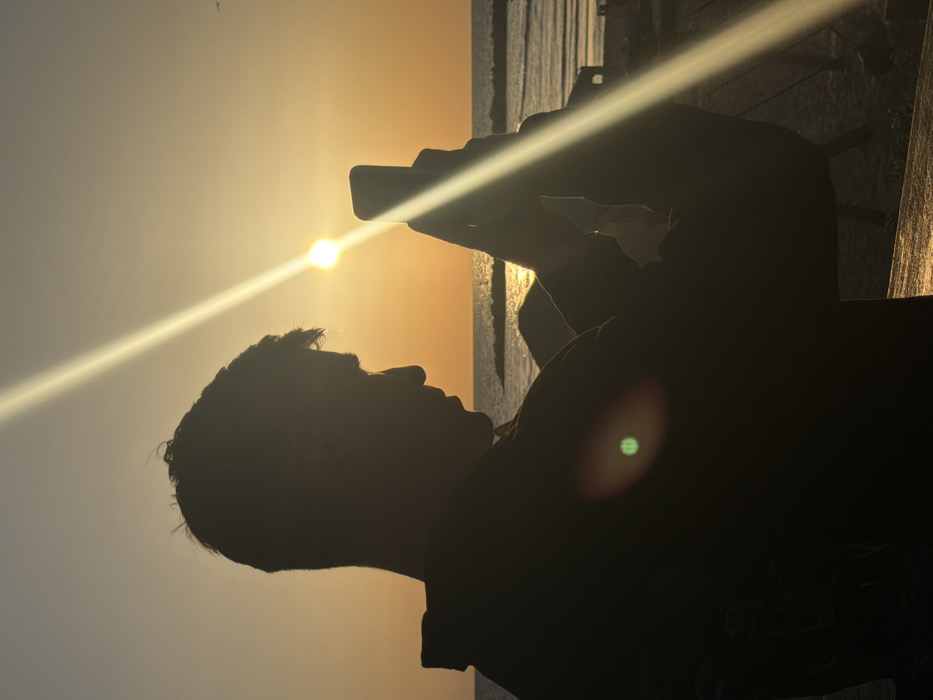 An individual, silhouetted and appearing male, is depicted in profile from the upper torso, facing right, at sunset on the Mediterranean coast in Tel-Aviv, Israel. The figure holds a smartphone vertically in their right hand, positioned as if capturing the scene. The sun, a distinct bright disk, is located near the horizon, slightly to the right of the figure's head, casting intense lens flares, including a prominent diagonal beam across the image and a green circular flare on the figure's back. The sky is illuminated with warm orange and yellow tones. In the background, the sea exhibits gentle waves, with dark structures resembling breakwaters visible in the water and faint outlines of structures on the sandy beach. The foreground elements are heavily underexposed, appearing as dark silhouettes against the bright sunset.