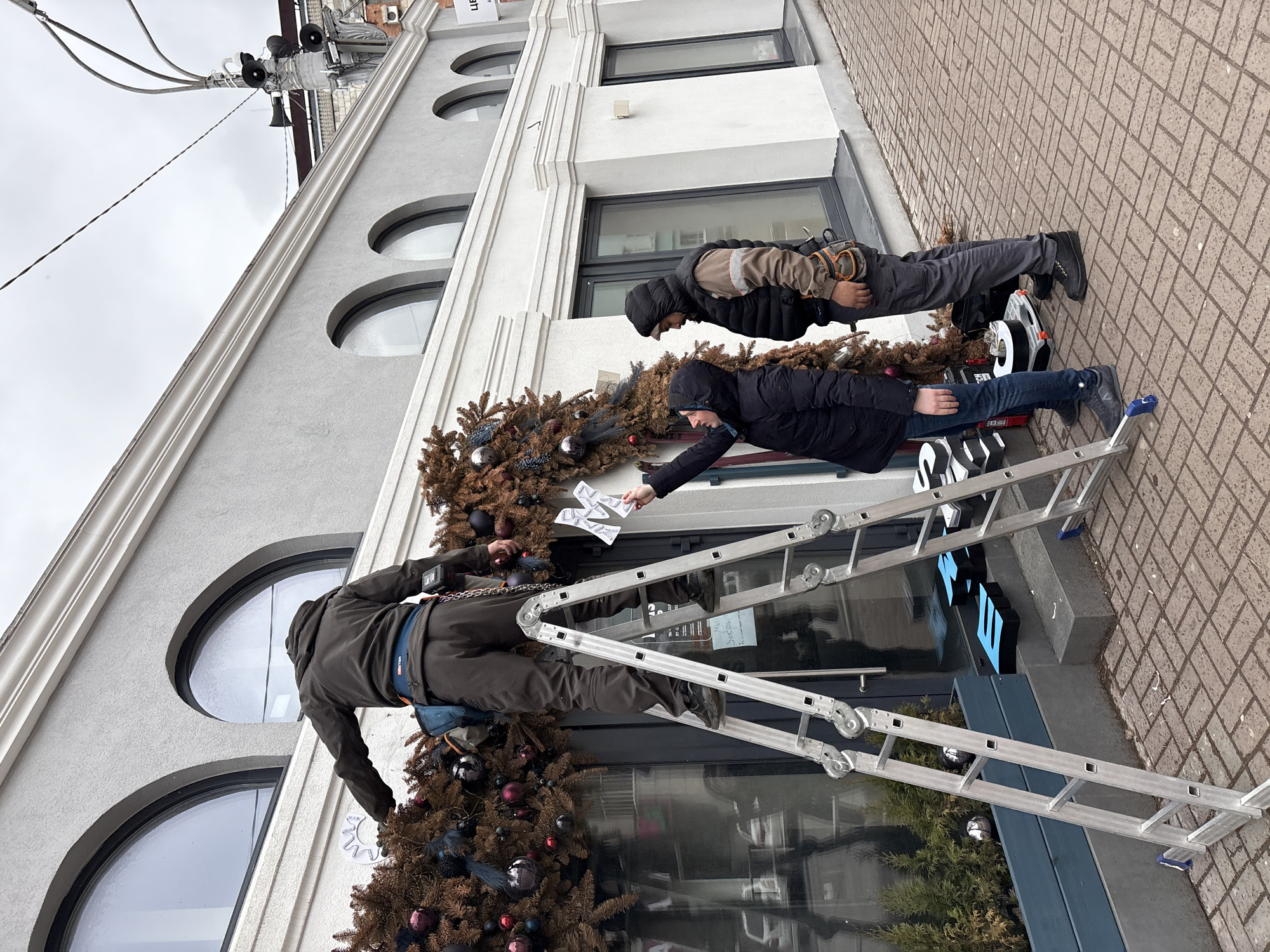 Three people are working together to decorate the exterior of a building. The building, of a grey and white color scheme, has large arched windows. Two men are on ladders, one near the top of the ladder is wearing brown pants and a brown jacket while attaching ornaments to a garland. The other man is standing on the building next to the first, he is wearing jeans and a black coat. A third person, a woman, is on the ground holding a set of papers that she is handing up to the person on the ladder, she is wearing a dark coat. The ladder is set on a brick walkway. From additional context clues, the location is in the city of Ulyanovsk, Russia.