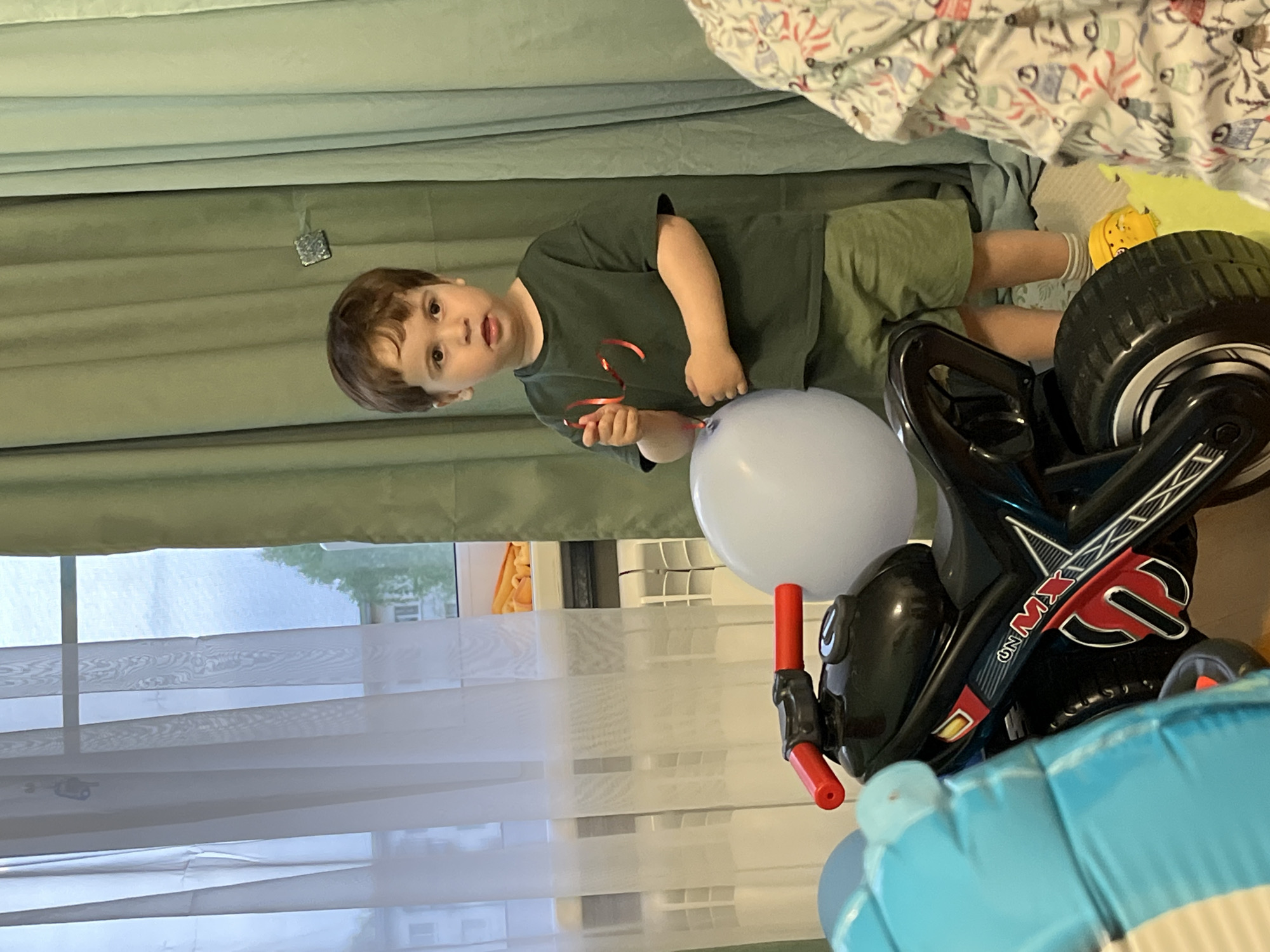 A young child in a green T-shirt and olive-green shorts stands indoors next to a toy motorcycle and a large light blue balloon. He holds the balloon’s string in his right hand. The motorcycle is black with red accents and has a prominent "ON MX" logo. The child wears yellow shoes with white socks. The background features a green curtain and a window with a white, patterned blind. Based on metadata, the image was taken in Chaikovsky, Russia.