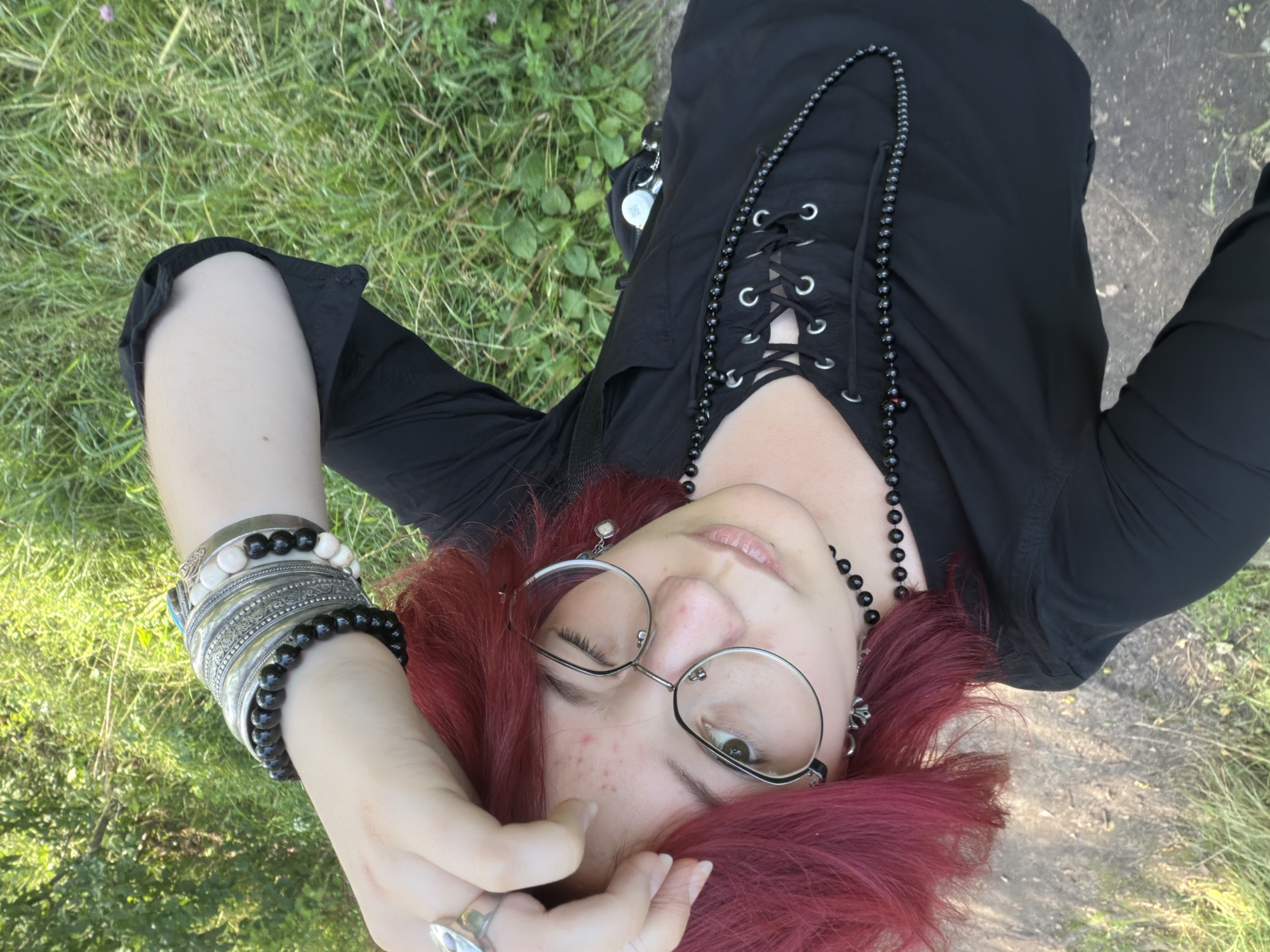 The image is a selfie taken outdoors, likely in a park or natural area. A person with vibrant red hair and glasses is looking towards the camera, with one eye winked shut. They are wearing a black, lace-up shirt and several necklaces and bracelets. The background consists of green grass and foliage, suggesting a sunny day. There is no indication of the city of Chelyabinsk, Russia, or any specific urban features.