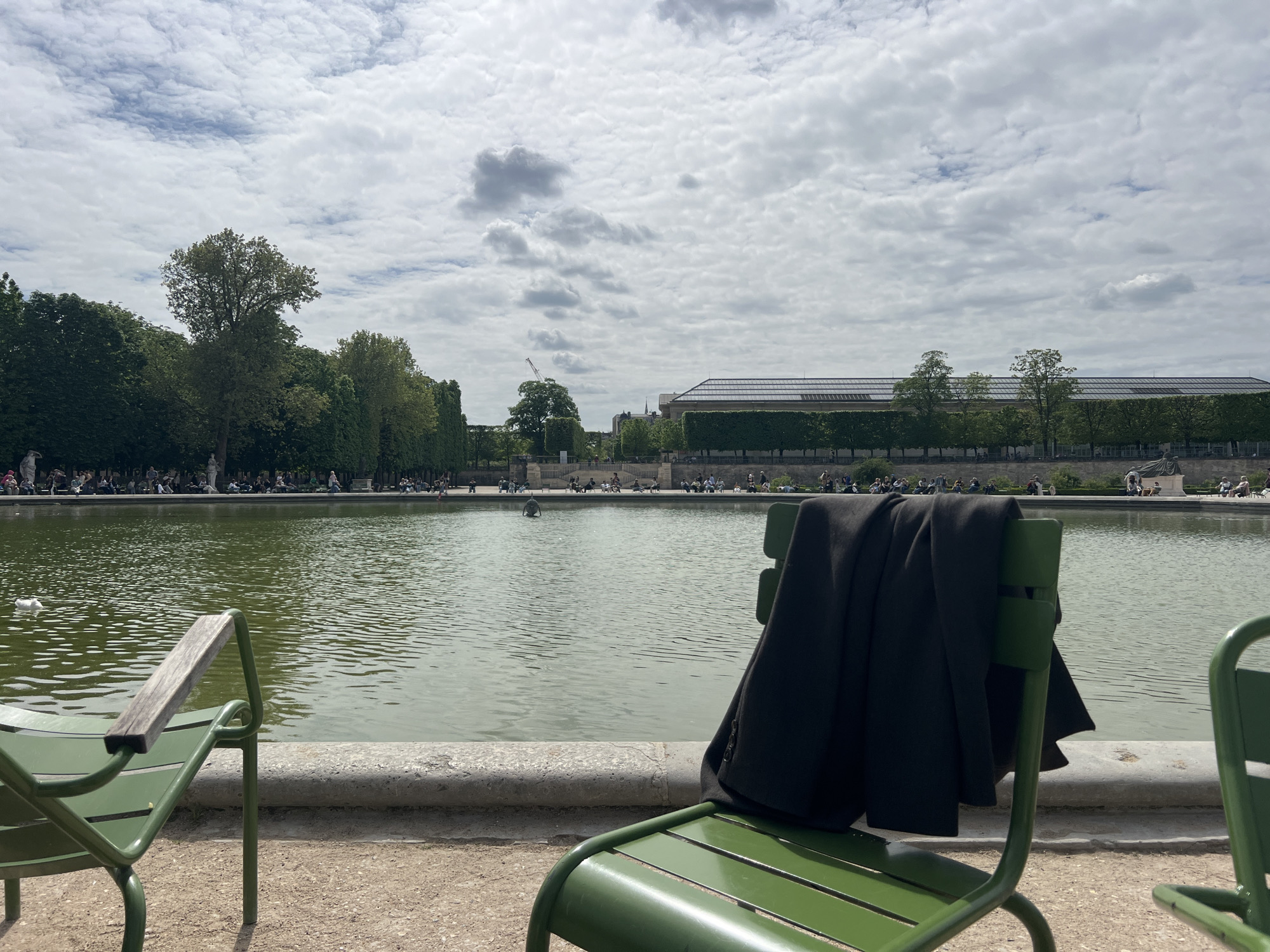 This image depicts a serene yet lively scene within a large public park, characteristic of Parisian gardens like the Tuileries or Luxembourg. In the foreground, two iconic green metal chairs are positioned, facing a large rectangular pond. A dark coat or jacket is casually draped over the back of the chair on the right, while the chair on the left has a wooden armrest.
The midground is dominated by the expansive pond, its surface gently rippling and reflecting the overcast sky. A white duck floats peacefully on the water on the left side.
Beyond the pond, numerous people are visible, scattered along a wide pathway and grassy areas. They are engaged in various leisure activities: sitting on benches, strolling, or simply relaxing by the water. A dense line of mature green trees forms a verdant backdrop behind the pathway.
In the far background, several buildings can be seen above the tree line. Some roofs feature modern elements like skylights or solar panels. A construction crane is faintly visible in the distance, poking above the trees.
The sky is filled with a mix of light and darker clouds, indicating an overcast or partly cloudy day, with diffused light suggesting it is daytime, possibly late morning or early afternoon. The overall mood is one of calm and leisure, with people enjoying the pleasant park environment. No discernible text is visible in the image.