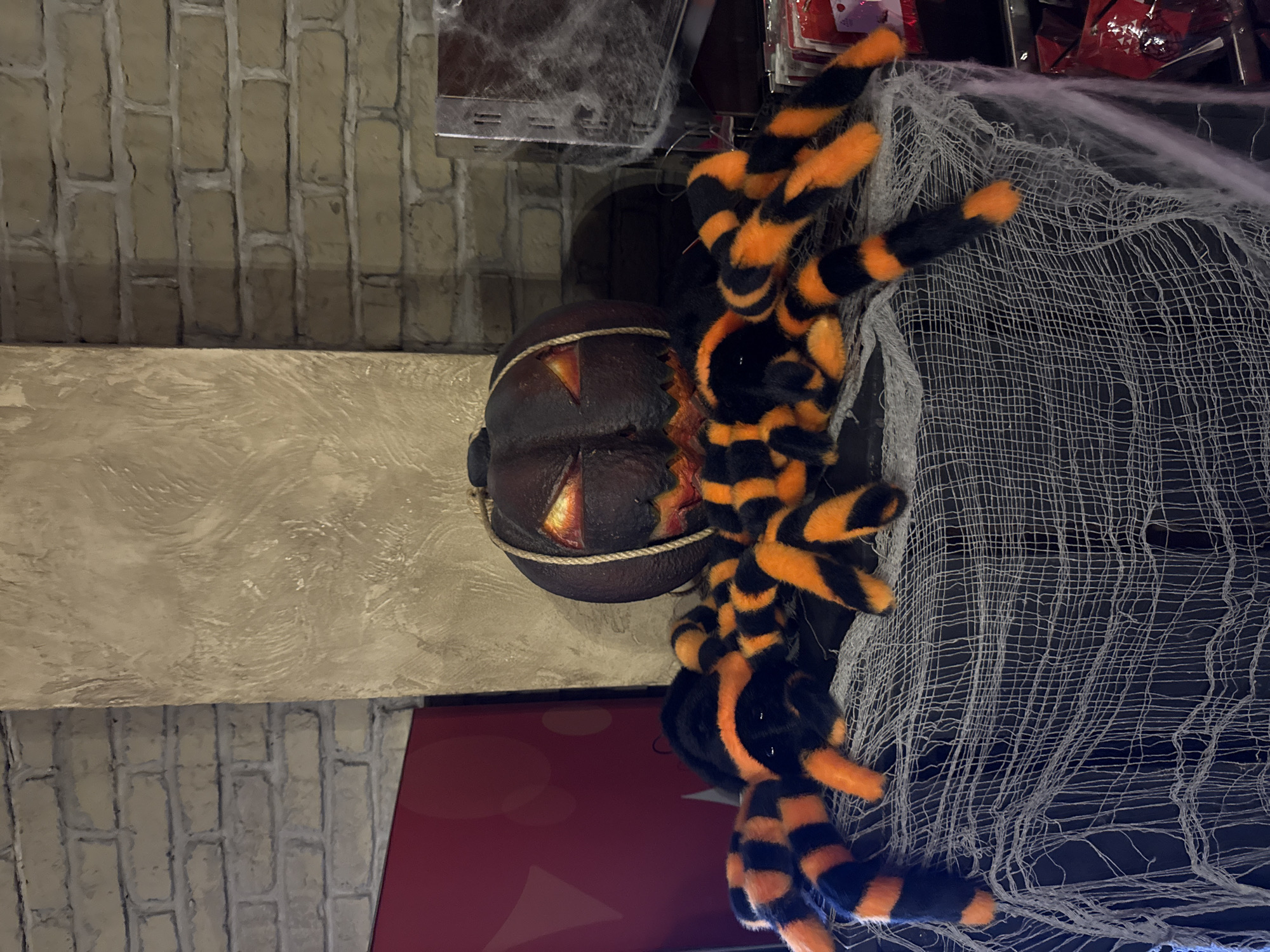 This image captures a Halloween-themed display in an indoor setting, likely a retail store or decorated space in Moscow, Russia. The scene is dominated by festive, spooky decorations.
A large, plush spider with distinct black and orange striped legs is a prominent subject, positioned to the right of the frame. Its numerous legs are splayed outwards, and it appears to be resting on or intertwined with white, semi-transparent faux spiderwebs, which are draped across a dark surface.
To the left of the spider, a dark, decorative pumpkin rests on a textured, light-brown beam or shelf that extends horizontally across the midground. The pumpkin is designed to look like aged wood or metal, wrapped with rope, and features carved, glowing eyes and a jagged mouth, resembling a jack-o'-lantern with internal illumination.
The background consists of a wall with a light-brown brick-like texture. Below the main beam, a glimpse of a darker surface and some blurry red and dark objects can be seen, possibly merchandise or storage, suggesting a commercial environment. The overall mood is festive and spooky, consistent with Halloween celebrations. There is no visible text in the image.