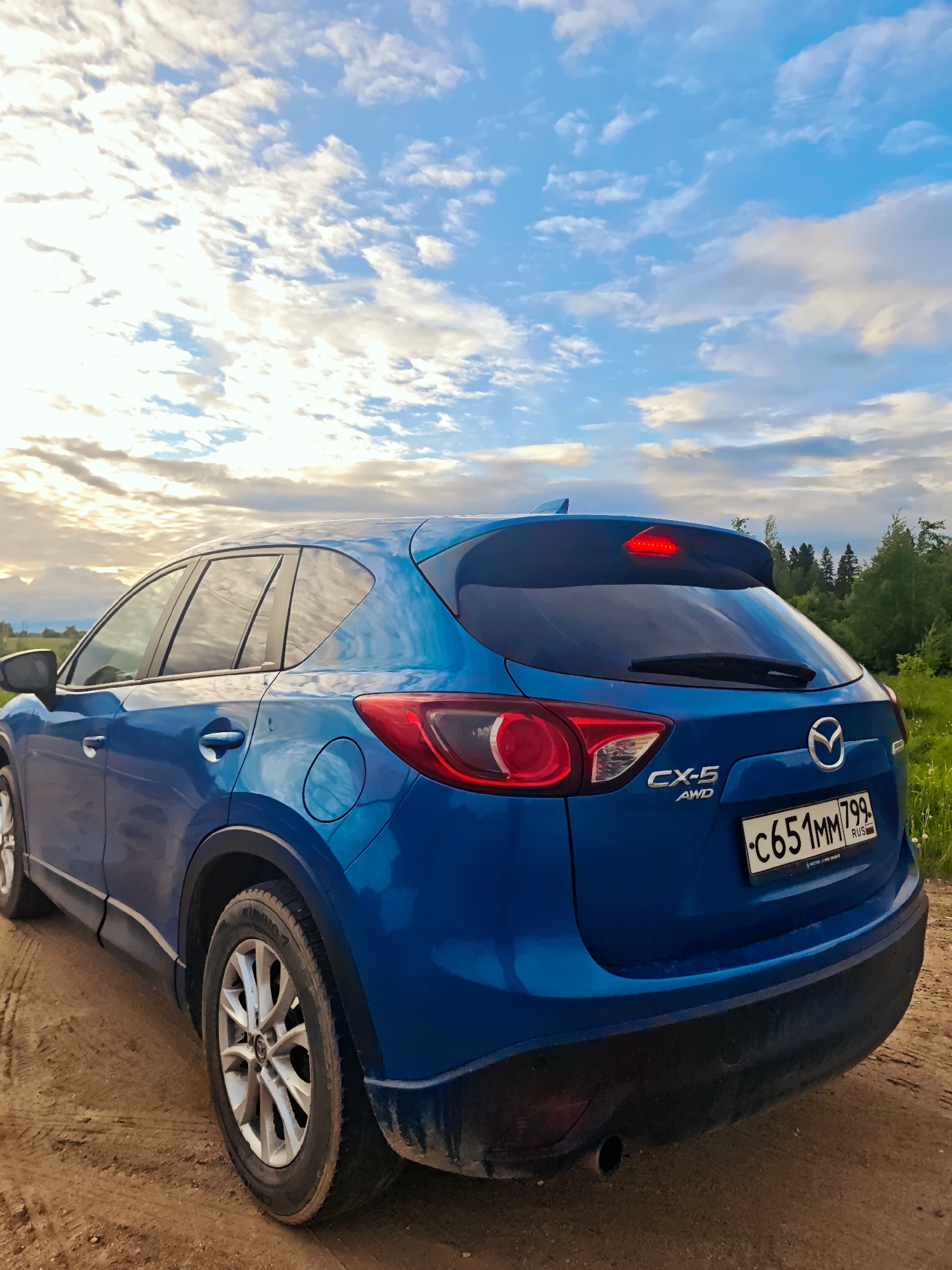 A blue Mazda CX-5 AWD is parked on a dirt road in what appears to be a rural or semi-rural area. The vehicle is facing away from the viewer and the rear passenger side is visible. The license plate reads "C651MM799 RUS". The background shows a field and trees, with a partly cloudy sky above. The scene is located in Курово, Russia.