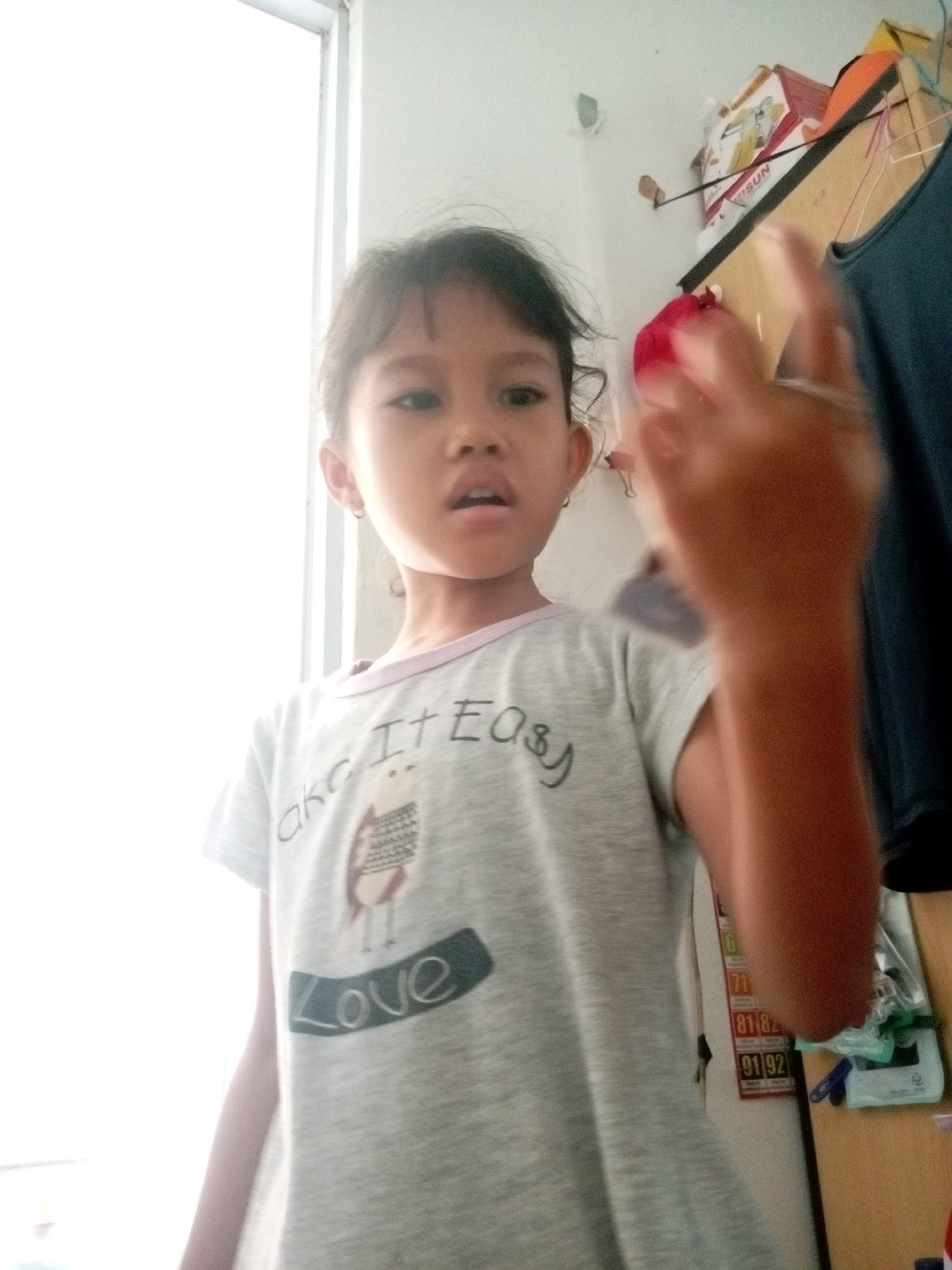 A young girl with dark hair, wearing a grey t-shirt with a graphic design that includes the word "Love," stands in what appears to be a room in Batam, Indonesia. She is looking towards the camera with her mouth slightly open. Her right arm is raised, and her hand is blurred in the foreground, holding a red object. Behind her, a wall is visible with various items, including boxes, what looks like a calendar with numbers, and some clothes hanging. A window with bright natural light is to her left, illuminating the scene.