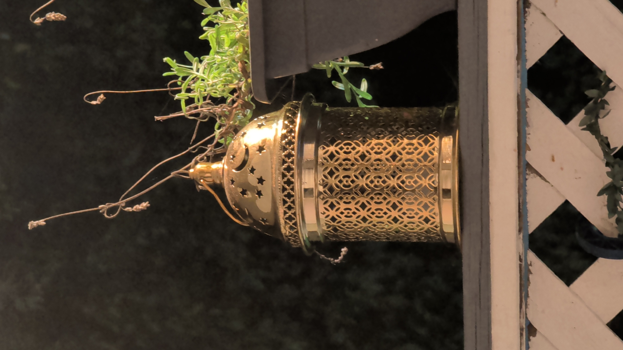 A brass lantern sits on a wooden railing. The lantern is cylindrical with a decorative filigree pattern and a domed top with star and crescent moon cutouts. A small loop of string hangs from the side of the lantern. Behind the lantern, a potted plant with green foliage is visible. The background is dark and out of focus. The railing is painted white with a distressed finish, revealing hints of blue underneath. The scene is lit from above, casting a warm glow on the lantern. There are no people or animals visible in the image. The environment appears to be outdoors, possibly a balcony or patio. The specific location in London, Canada, cannot be determined from the visual information provided.