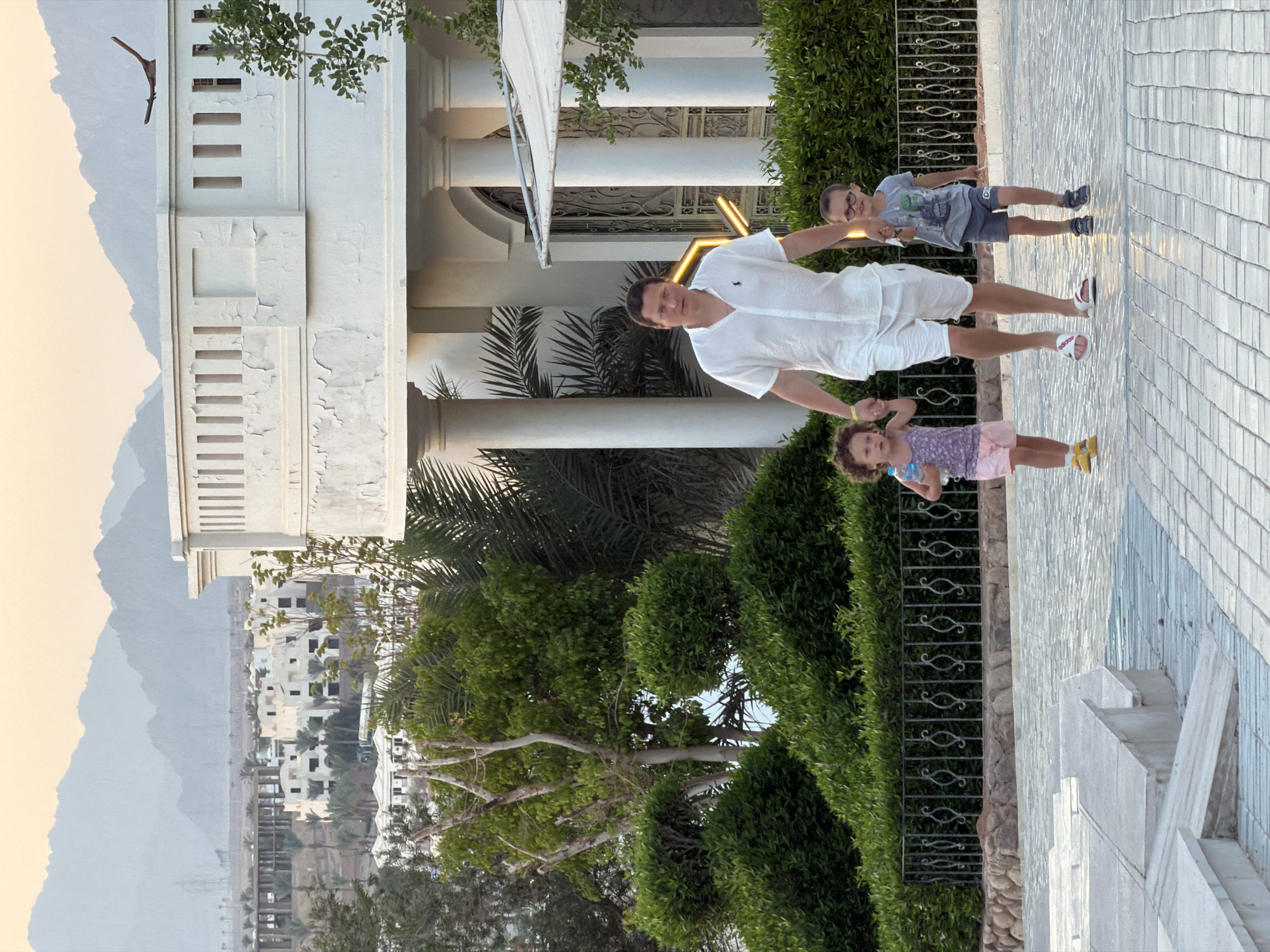 In Sharm El Sheikh, Egypt, a man in a white short-sleeved shirt and white shorts, wearing white sandals, walks with two children. The girl on the left, wearing a purple and white patterned sleeveless top and pink shorts with yellow shoes, holds a water bottle. The boy on the right, wearing glasses, a grey t-shirt with a graphic, and grey shorts with dark shoes, holds the man's hand. They are on a paved walkway in front of a lush green hedge. Behind them, a white building with columns and decorative elements is visible, and in the distance, hazy mountains can be seen under a pale sky.