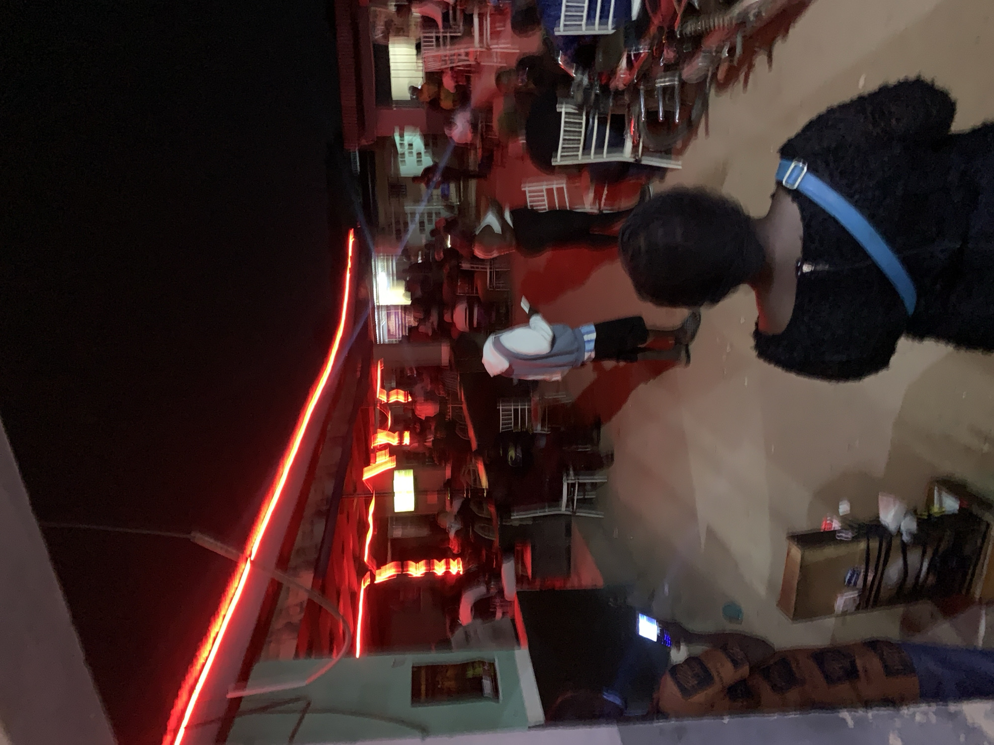 The scene is a nighttime gathering in Kabong, Nigeria. People are seated at tables and chairs, likely at an outdoor event, potentially a bar or restaurant. Red neon lights illuminate a building facade. A person wearing a black jacket with a blue strap is visible in the foreground. Other individuals are present but their features are not distinct. A building with lit windows is partially visible.