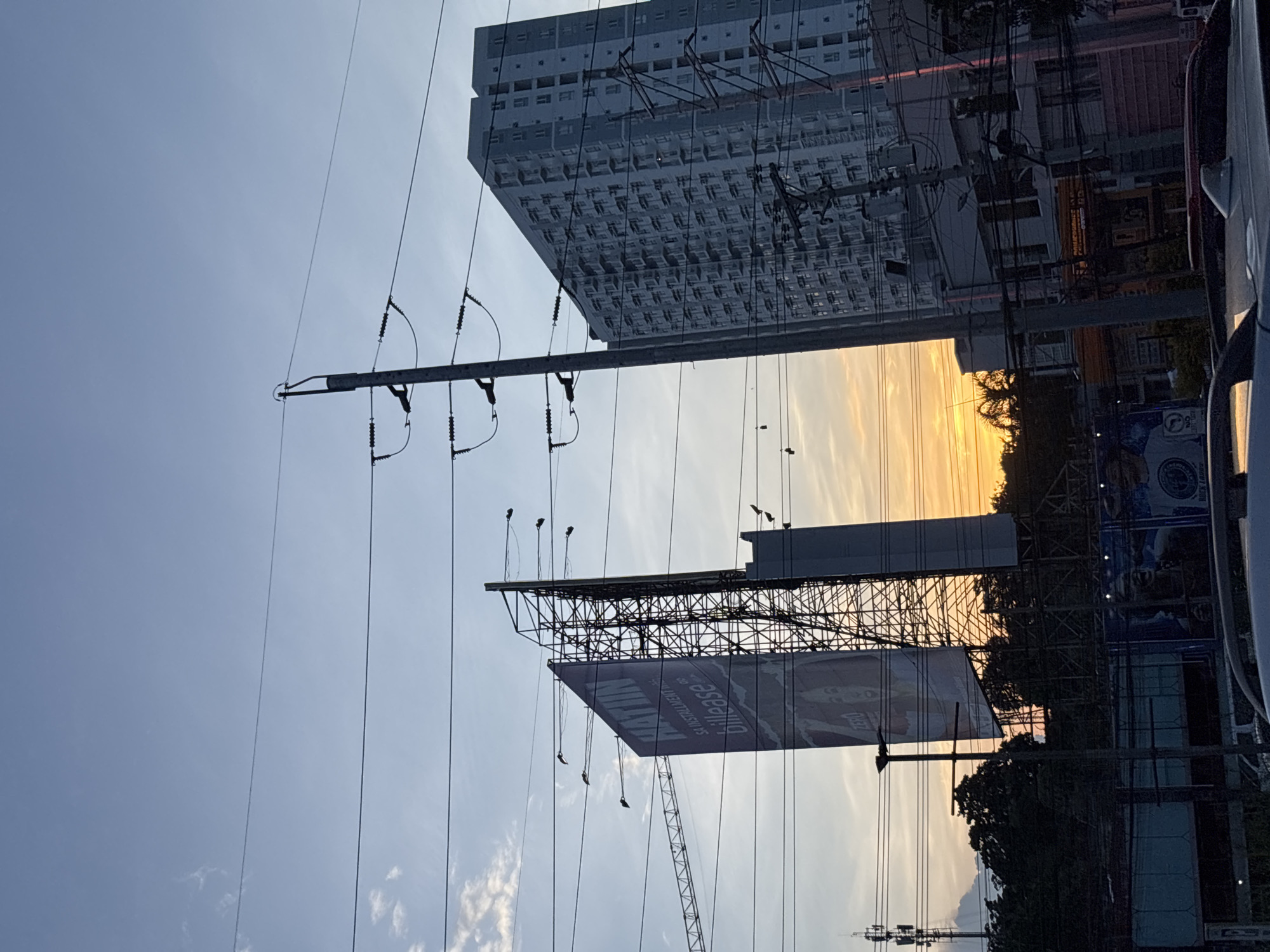 The image depicts a street-level view of an urban scene at dusk in Davao City, Philippines. Dominating the composition are power lines and a large billboard advertising "HATIN sa installment with billeease," featuring a woman's face. To the right, a tall, modern residential building with many windows stands out. In the background, the sky is a soft blend of blue and orange, indicating the setting sun, with scattered clouds. Silhouetted against the horizon are trees and other structures, including a cell tower on the left. Several cars are partially visible in the foreground. The overall atmosphere is that of a typical city evening, with infrastructure and advertising elements prominently displayed.
