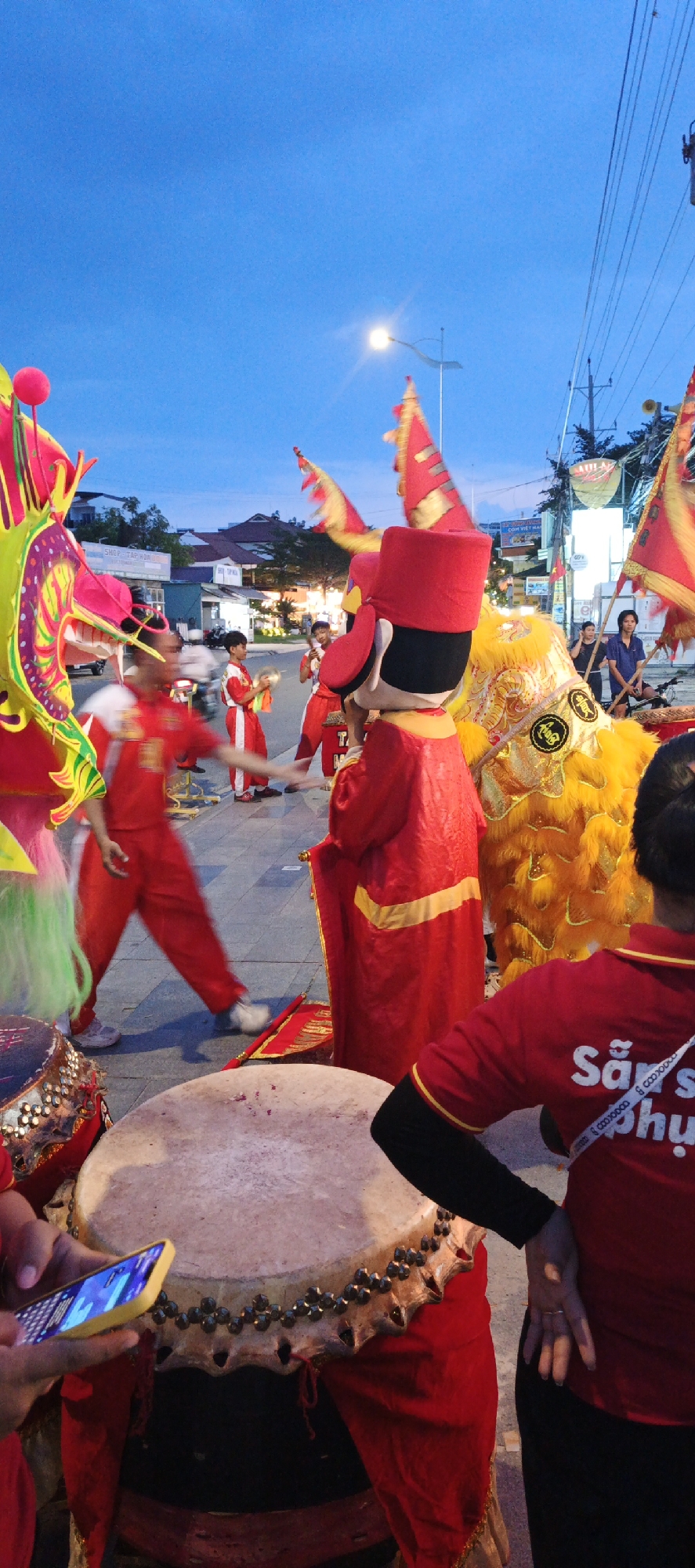 The image depicts an outdoor cultural performance taking place at dusk on a paved street in an urban environment.
In the immediate foreground, two large traditional drums are visible. On the left, a hand holding a bright yellow-cased smartphone is positioned above the drumhead, with the phone's screen illuminated. To the right, a person is seen from the back, wearing a red short-sleeved shirt with the white text "Sản phụ" on it, a black long-sleeved undershirt, and black pants.
The mid-ground is dominated by performers and their elaborate costumes. On the far left, a person is partially visible, operating a brightly colored dragon head, featuring prominent green, yellow, and pink scales. Central to the frame, a performer is dressed in a full red costume with a distinctive tall, cylindrical red hat. To their right, a vibrant yellow lion dance costume is present, adorned with black Chinese characters, notably "福" (fú), on its head and body. Several young male performers in matching red tracksuits are also observed; one on the left holds a pair of cymbals, while others are in various stages of movement, some appearing blurred. Red and gold flags on poles are positioned behind these performers.
The background reveals a street lined with buildings. A white sign with blue text, "SHOP TẬP HỌC M...", is visible on one building, and another sign further down reads "Cơm Việt Nam". A blurred motorcycle is captured mid-passage, and a bicycle is parked near onlookers on the right side of the street.
The upper section of the image features a deep blue sky, indicative of dusk or early evening. A prominent, brightly glowing street lamp is positioned in the upper center-right, casting light onto the scene. Overhead power lines are visible traversing the sky.