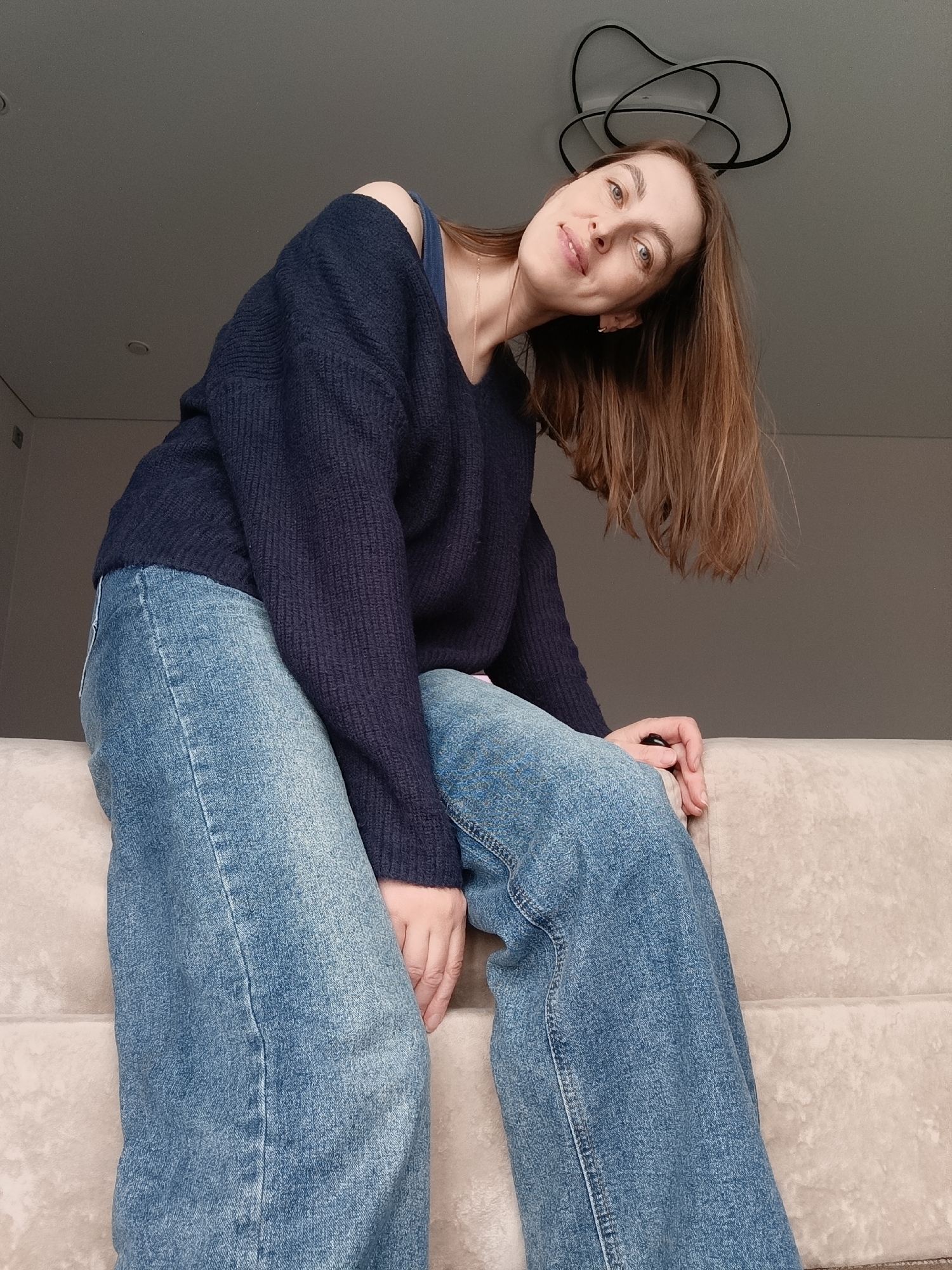 A woman with light skin and brown hair, wearing a navy blue sweater and blue jeans, sits on a light beige upholstered sofa or couch. She is leaning forward and slightly to her left, looking at the camera with a small smile. Her head is tilted to the side. Part of her shoulder is exposed. A silver necklace is visible on her neck. The room has neutral-colored walls and modern lighting fixtures. The location is Chaikovsky, Russia.