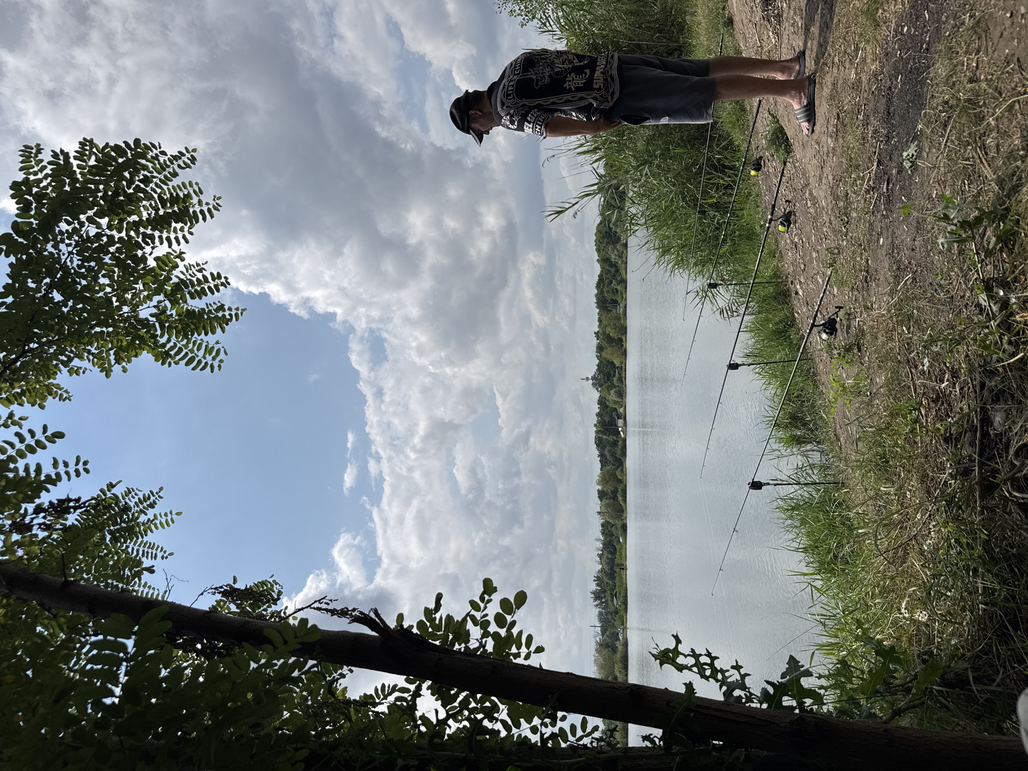 A man stands on the bank of a body of water, facing away from the camera and looking out at the water. He is wearing a black baseball cap, a black and white patterned short-sleeved shirt, and grey shorts. Several fishing rods are set up in the foreground, some propped on stands. The man is standing near tall green reeds. The water is calm and reflects the sky, which is partially cloudy. Trees line the opposite bank of the water. The scene appears to be a natural outdoor environment, possibly a lake or river, located in Kochubeyevskoye, Russia.