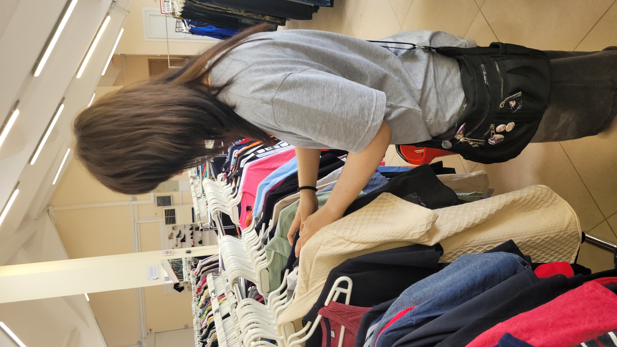 A medium shot depicts a person with long, dark brown hair leaning over a clothing rack in what appears to be a store. The person is wearing a light gray t-shirt and a black bag with pins on it. The clothing rack is filled with clothes in various colors, hung on white plastic hangers. The environment seems to be a retail setting, with bright overhead fluorescent lighting. The location is given as the city of Unknown, Unknown.