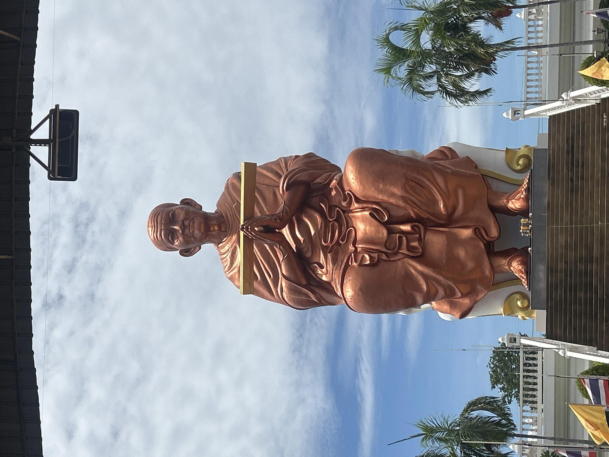 A large, bronze statue of a Buddhist monk is depicted sitting cross-legged in front of a cloudy blue sky. The statue is positioned on a raised platform accessible by a wide set of stairs. The monk statue is wearing traditional robes and has its hands clasped together at chest level, holding a horizontal wooden beam. The statue has a serene expression. In the foreground, on the left side, are flags of Thailand and possibly other national flags. Palm trees are visible on either side of the statue, adding to a tropical atmosphere. The overall setting suggests a religious or cultural landmark in a sunny, outdoor environment. The location is identified as Thai Ko Subdistrict, Thailand.