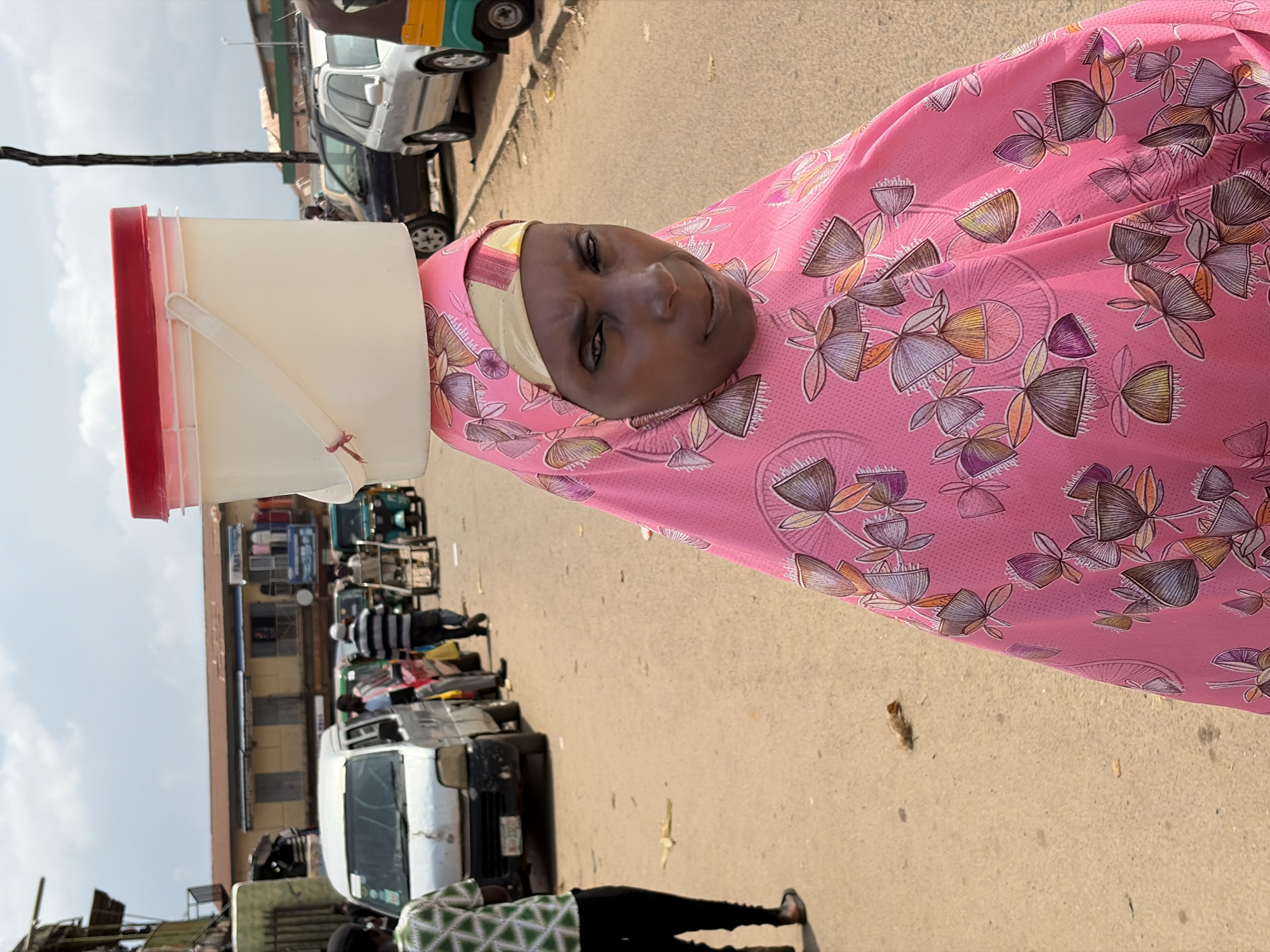 A woman walks on a paved road in Jos, Nigeria, balancing a large white plastic bucket with a red lid on her head. She is wearing a pink headscarf and a matching pink garment with a floral pattern. In the background, a white van, a black car, a silver car, and a green motorized tricycle are parked along the road. Buildings line the street, with some appearing to be shops or market stalls. The sky is cloudy.