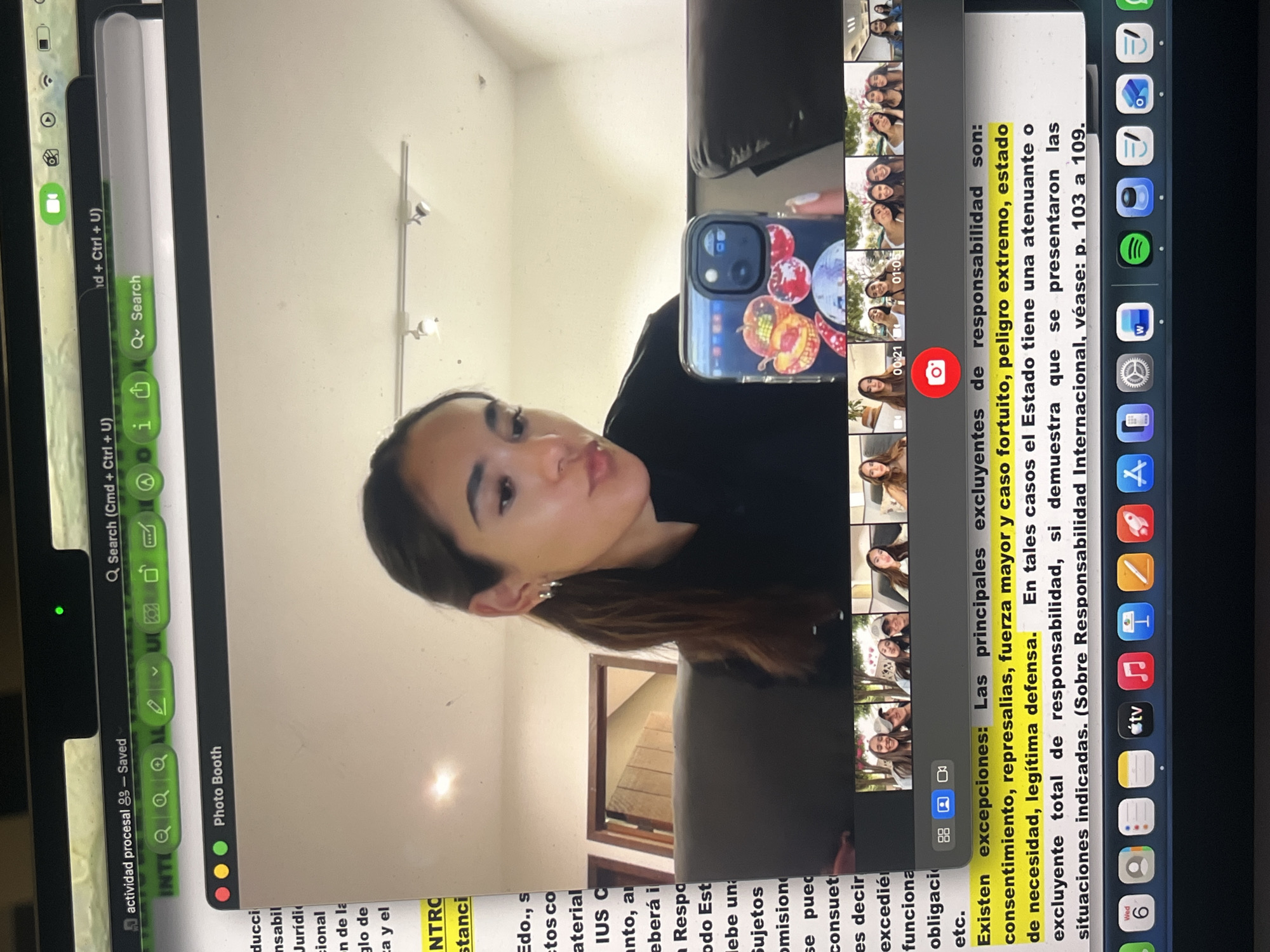A woman is using a smartphone to photograph a computer screen. The computer screen displays a video conferencing application showing multiple thumbnail videos of the woman in different poses. The computer also shows other open applications, including a document with highlighted text in Spanish about exceptions to responsibility, legal in nature, as well as a Photo Booth application. Various application icons are visible on the computer's dock. The background suggests an interior space, possibly a home. The location, San Bernardino Tlaxcalancingo, Mexico, is not discernible from the image provided.