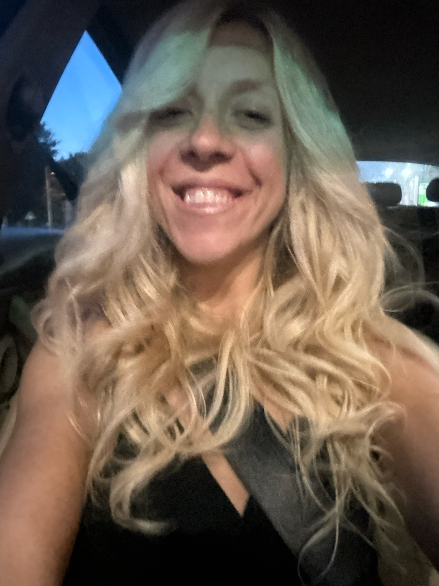 A woman with blonde, wavy hair smiles at the camera while sitting in the driver's seat of a car. She is wearing a black tank top and a gray seatbelt is visible. The sky is visible through the car's windshield, with hints of trees and streetlights suggesting an urban environment, possibly Sofia, Bulgaria.