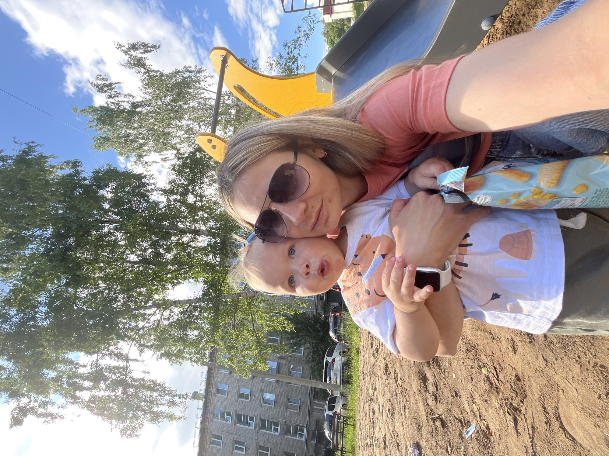 A woman with blonde hair and sunglasses is taking a selfie on a playground with a child she is holding. The child, who appears to be a toddler, is looking at the camera with an open mouth. The woman is wearing a coral-colored t-shirt and jeans, while the child is wearing a white t-shirt with an animal print and olive-green shorts. The child also has a smartwatch on their wrist. In the background, there is a yellow and gray slide, trees, a multi-story apartment building, and parked cars. The ground is covered in sand. The image was taken in Chaikovsky, Russia.