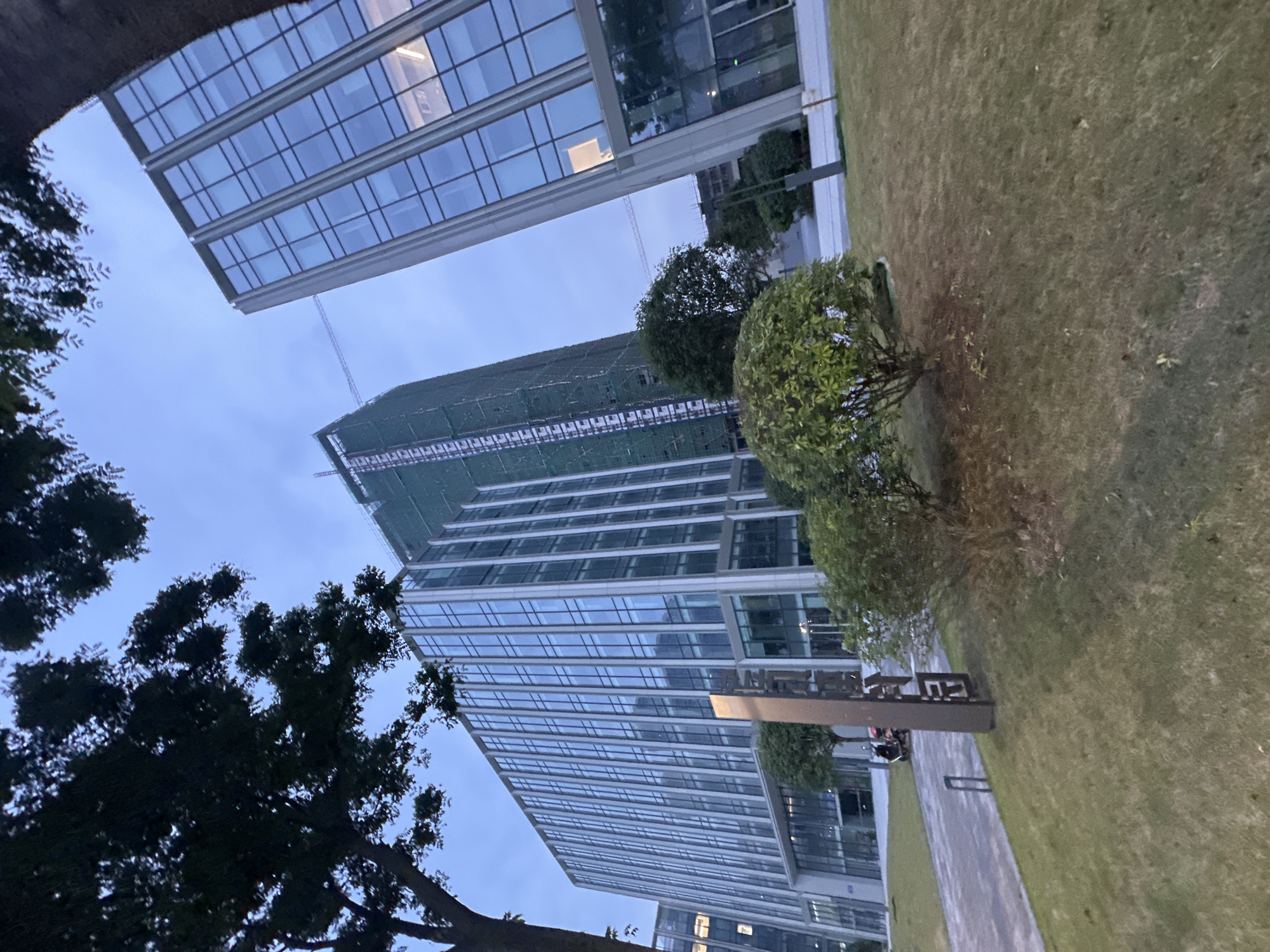 The image captures a contemporary architectural scene in the Chongqing High-tech Zone, China, under an overcast sky. The foreground features a manicured lawn with two rounded bushes. A dark, vertical monument or sign with stylized Chinese characters is prominently placed on the grass. To the left, a paved walkway leads towards a modern glass-fronted office building. A scooter is parked near the walkway, and two people are seated on a bench in the distance.
The primary focus is on the surrounding architecture. A long, multi-story office building with a reflective glass facade dominates the left and center of the frame. Its structure is characterized by vertical white mullions dividing numerous glass panels. To the right, another, taller glass-fronted building is angled towards the viewer. This building also displays a grid-like pattern of glass and white framing. Behind the first office building, a construction site is visible, with a taller structure partially covered in green scaffolding and netting, suggesting ongoing development. A construction crane is also visible in the background between the two glass buildings. Trees with dark foliage frame the top of the image, their branches reaching into the sky. The overall impression is one of a modern business district with ongoing construction and landscaping.