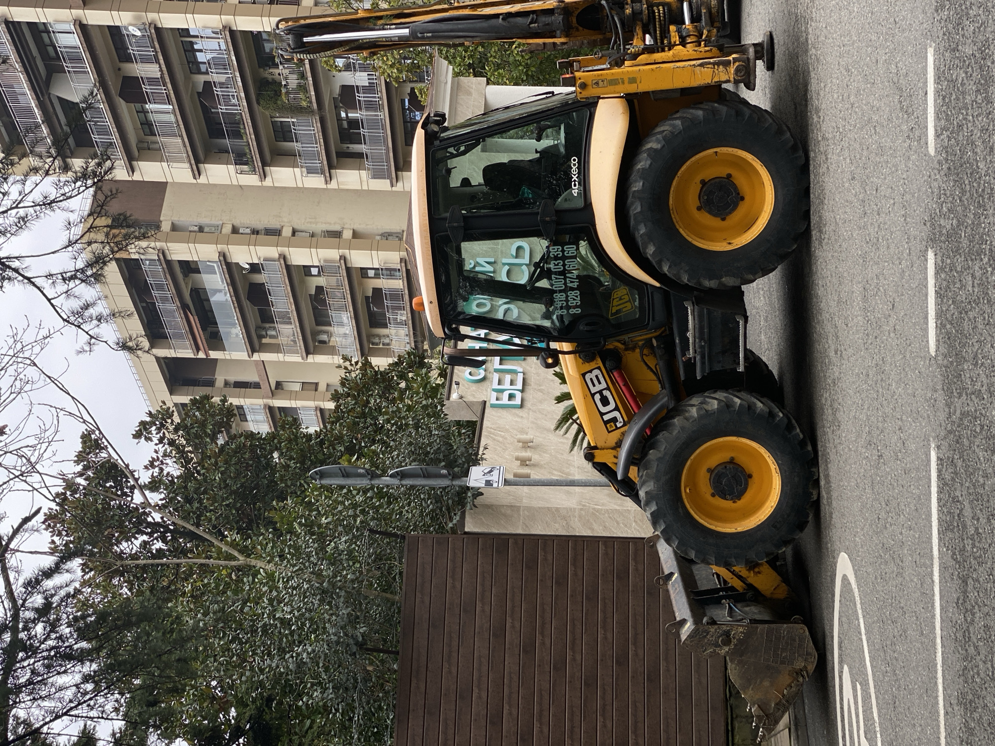 A yellow JCB 4CXECO backhoe loader is positioned on an asphalt road under an overcast sky. The vehicle faces slightly left, with its front loader bucket lowered to the pavement. A person is visible in the operator's cab. The right side of the vehicle displays the text "JCB" and "4cxeco," along with two phone numbers: "8 918 007 03 39" and "8 928 474 60 60." White road markings are present on the asphalt.
To the left of the vehicle, a dark brown, slatted fence stands adjacent to a light-colored building wall. A street light pole with a 'tow away' sign is visible. The wall behind the vehicle features green lettering that reads "САНАТОРИЙ БЕЛАРУСЬ" (Sanatorium Belarus). This signage indicates the location as the Sanatorium Belarus in Sochi, Russia, specifically at Kirova Street, 44.
Behind the Sanatorium Belarus, multi-story buildings with numerous balconies, some with glass railings and others with metal, are visible. These buildings exhibit various shades of beige and brown on their facades and feature multiple external air conditioning units and satellite dishes. Lush green trees and shrubs are scattered throughout the scene, with some bare tree branches overhead in the foreground.