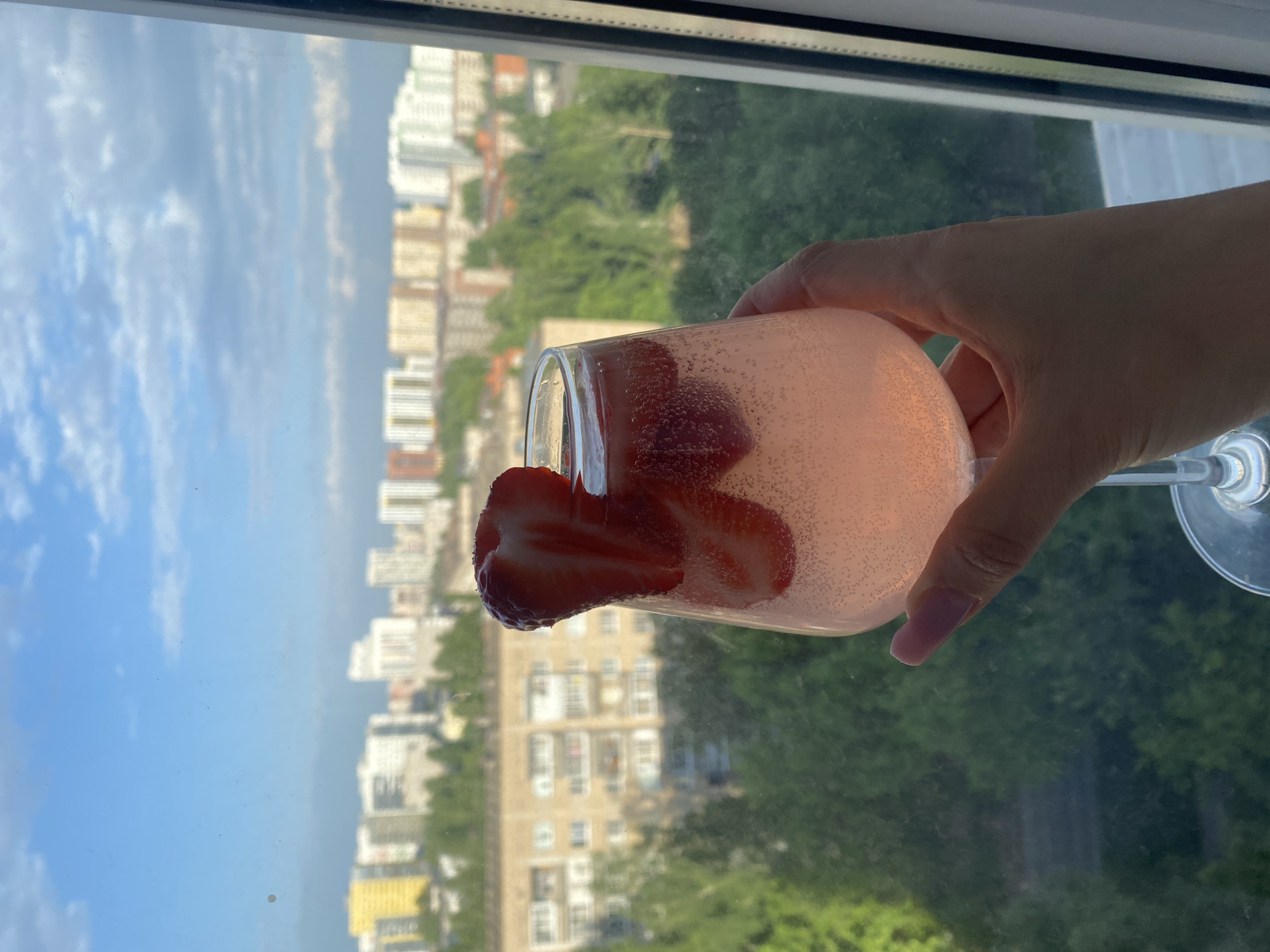 A person is holding a glass of a pink beverage, likely sparkling wine or soda, with sliced strawberries in front of a window. The background shows a cityscape with multiple apartment buildings and trees. The location is Izhevsk, Russia.